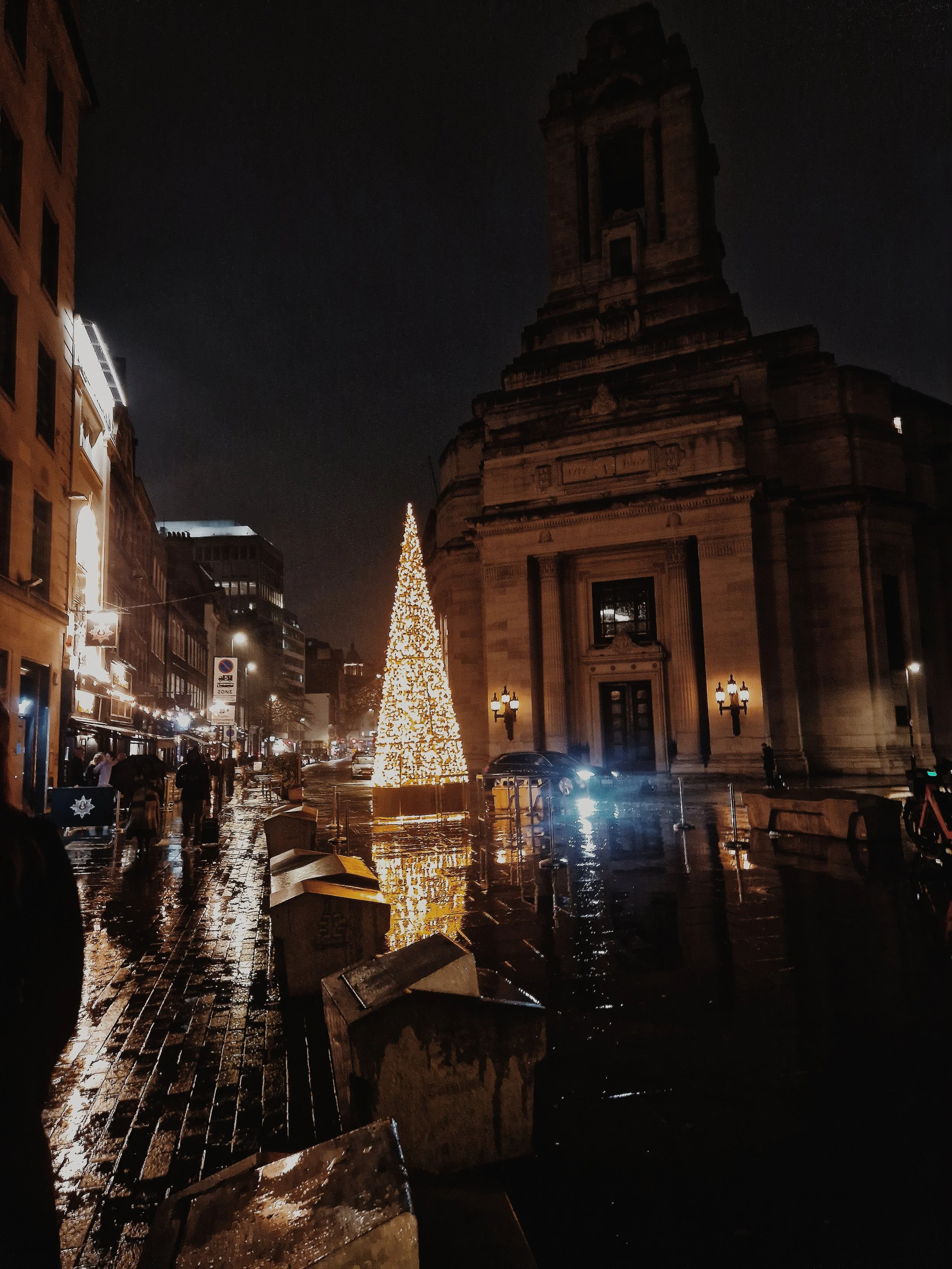 Night scene of a city street with a Christmas tree lit up with white lights, a large historical building, wet cobblestone pavement reflecting the lights, and a few people walking with umbrellas.