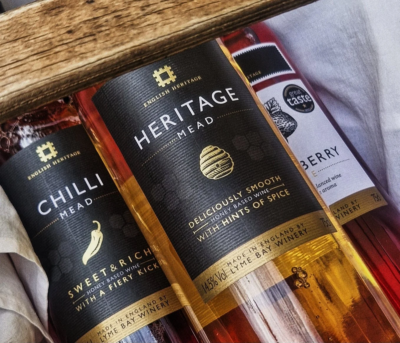 Three bottles of flavored mead lying on their sides inside a wooden box. The bottles are labeled 'Heritage Mead' with flavors 'Chilli' and 'Smooth' in the foreground and 'Berry' slightly behind.