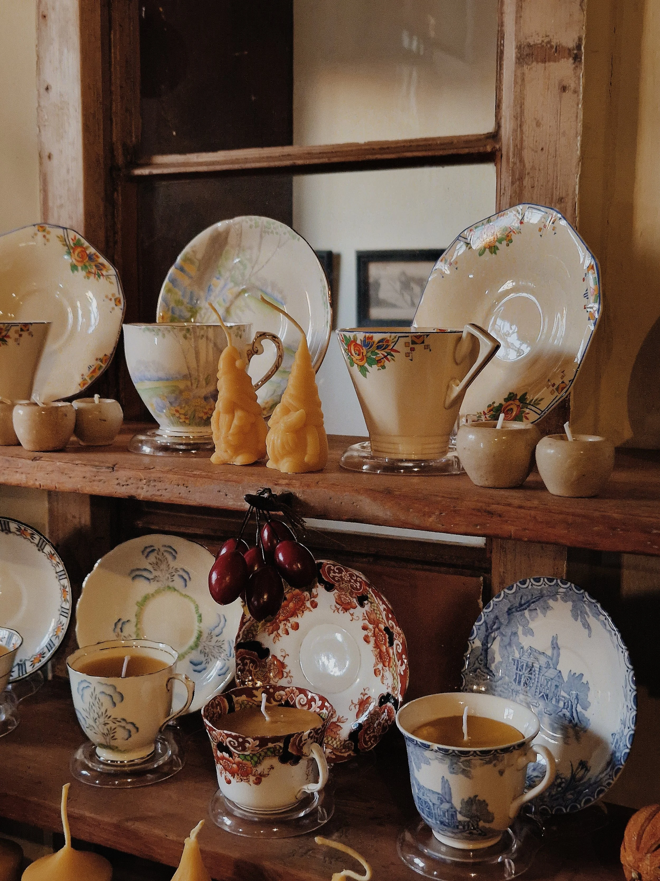 Display of vintage china tea cups and saucers, decorative beeswax candles, and a bunch of dark red grapes on a wooden shelf.