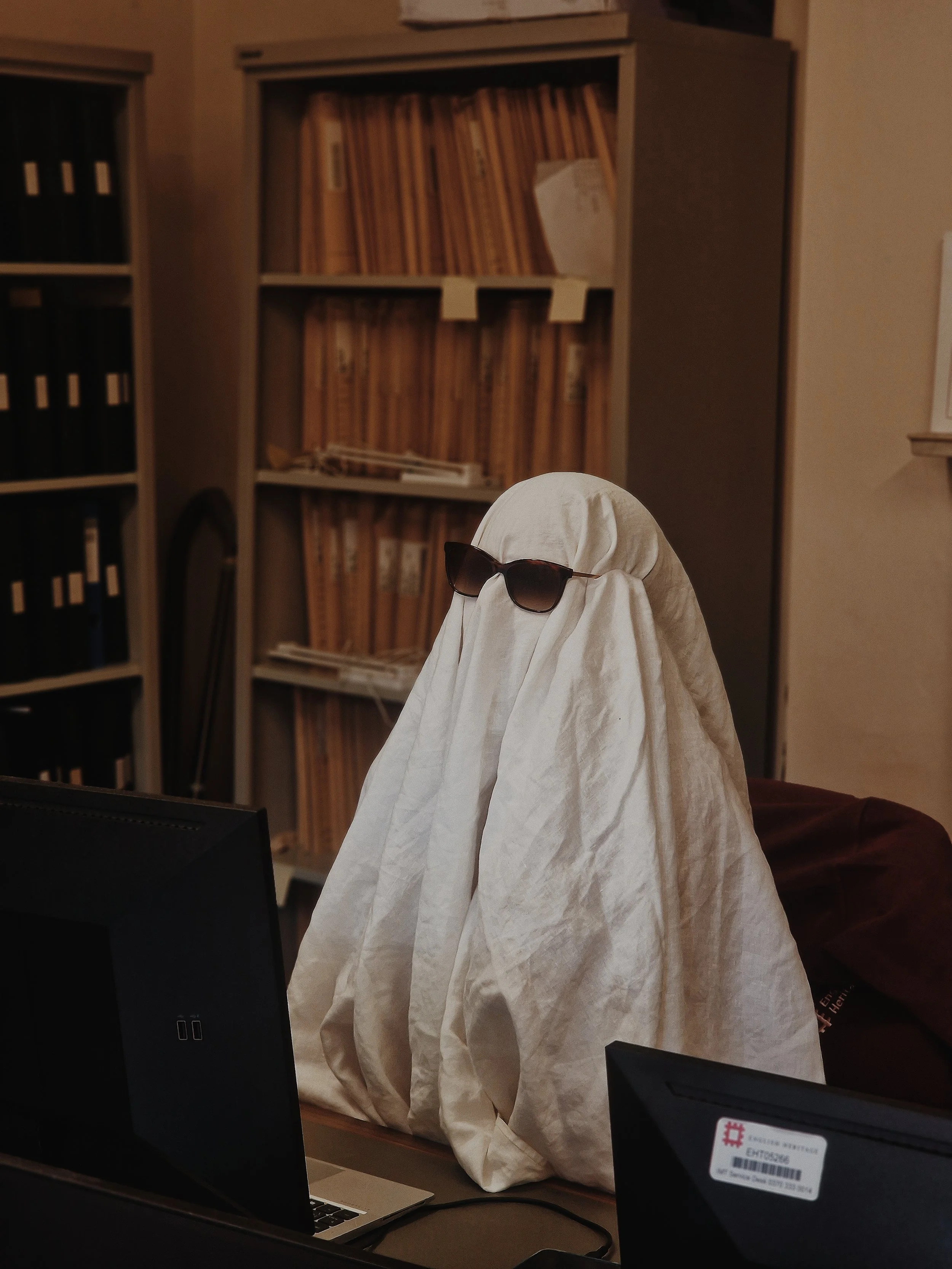 A figure draped in a white sheet with black sunglasses sitting in front of a computer in an office, with shelves of files in the background.
