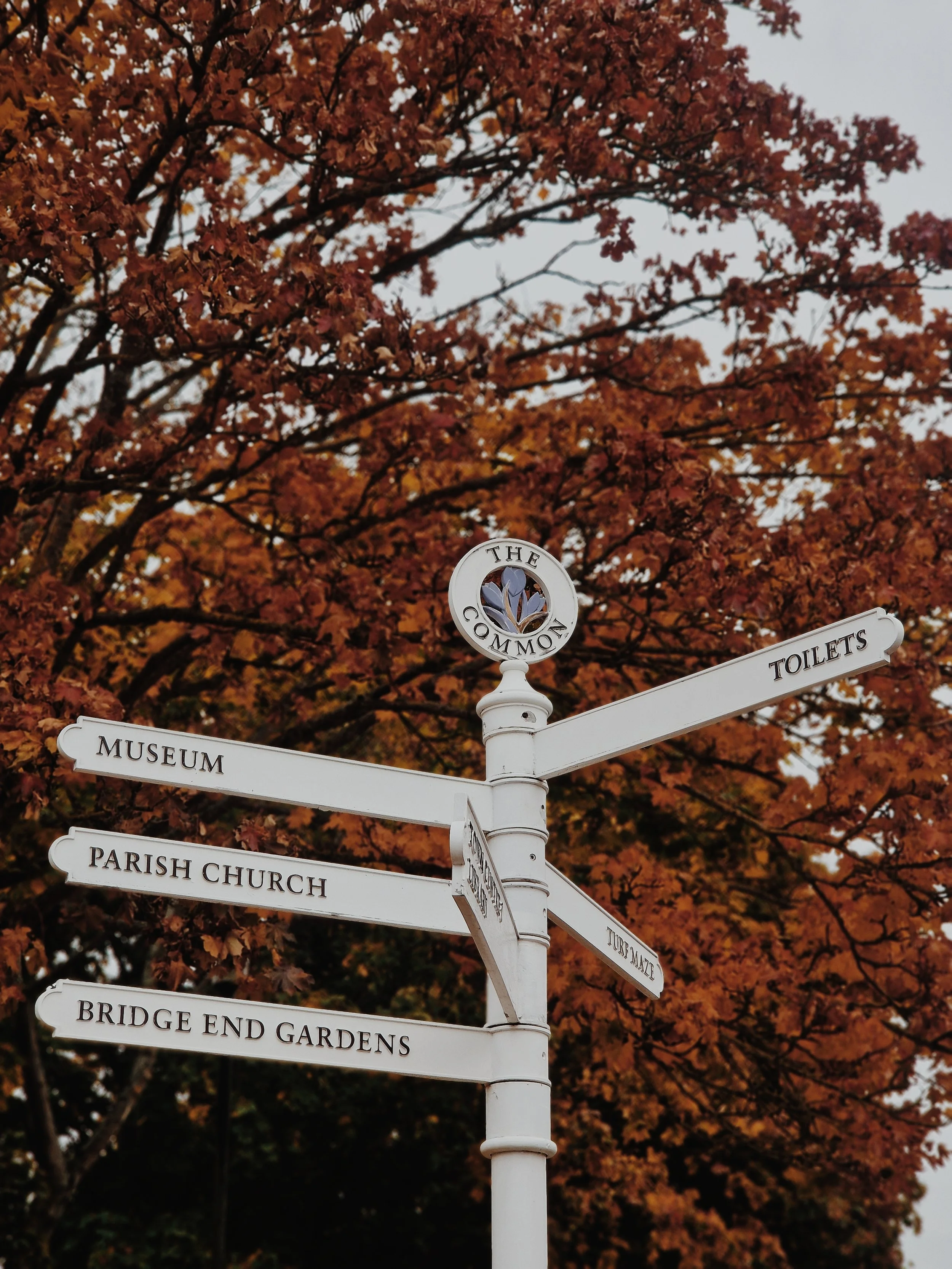 White signpost with multiple directional signs, including museums, a parish church, a garden, a pub, and toilets, set against a background of orange autumn leaves on a tree.