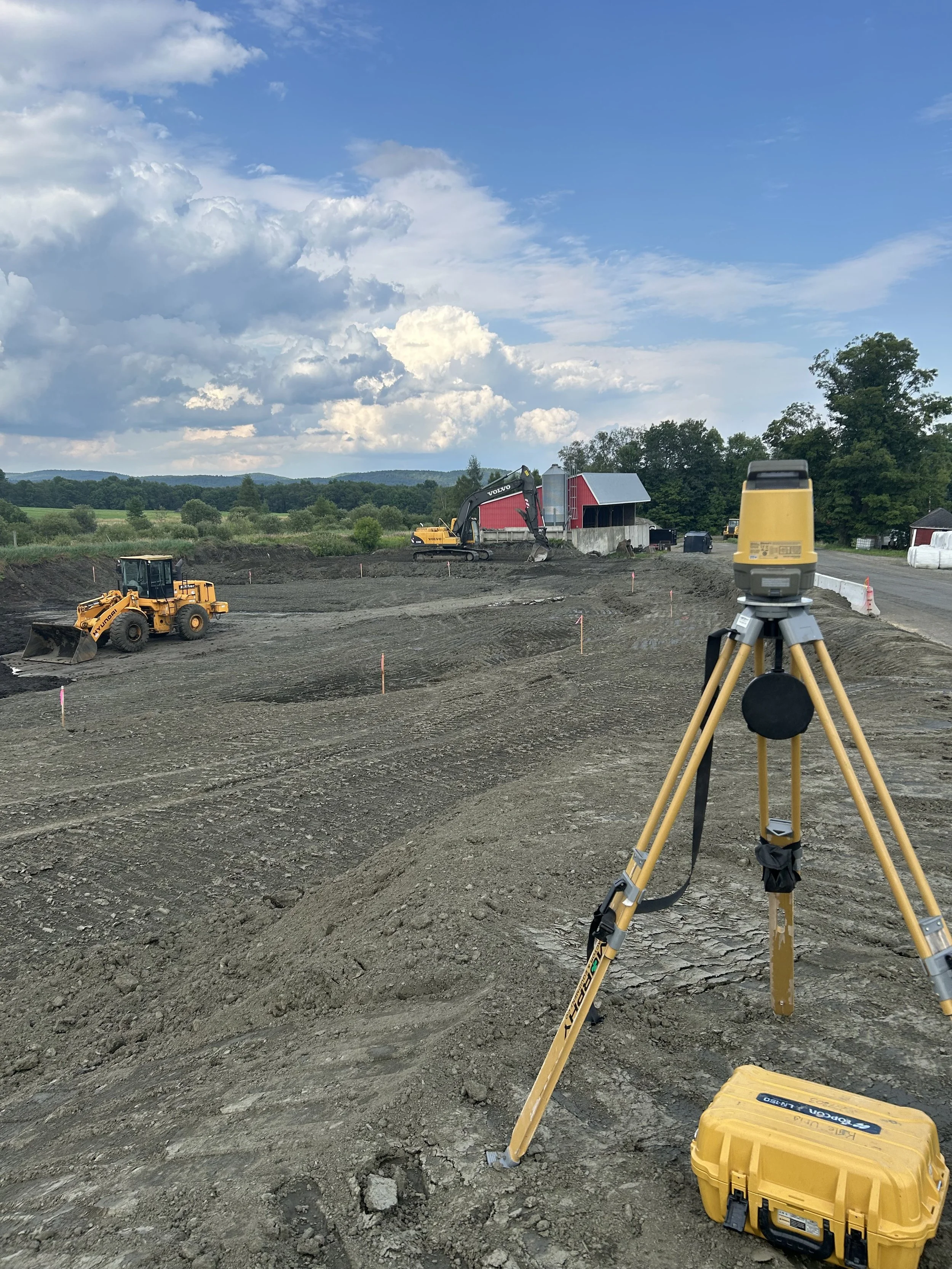Construction site with earth-moving equipment, a surveyor's level on tripod, and a yellow toolbox, under a partly cloudy sky.