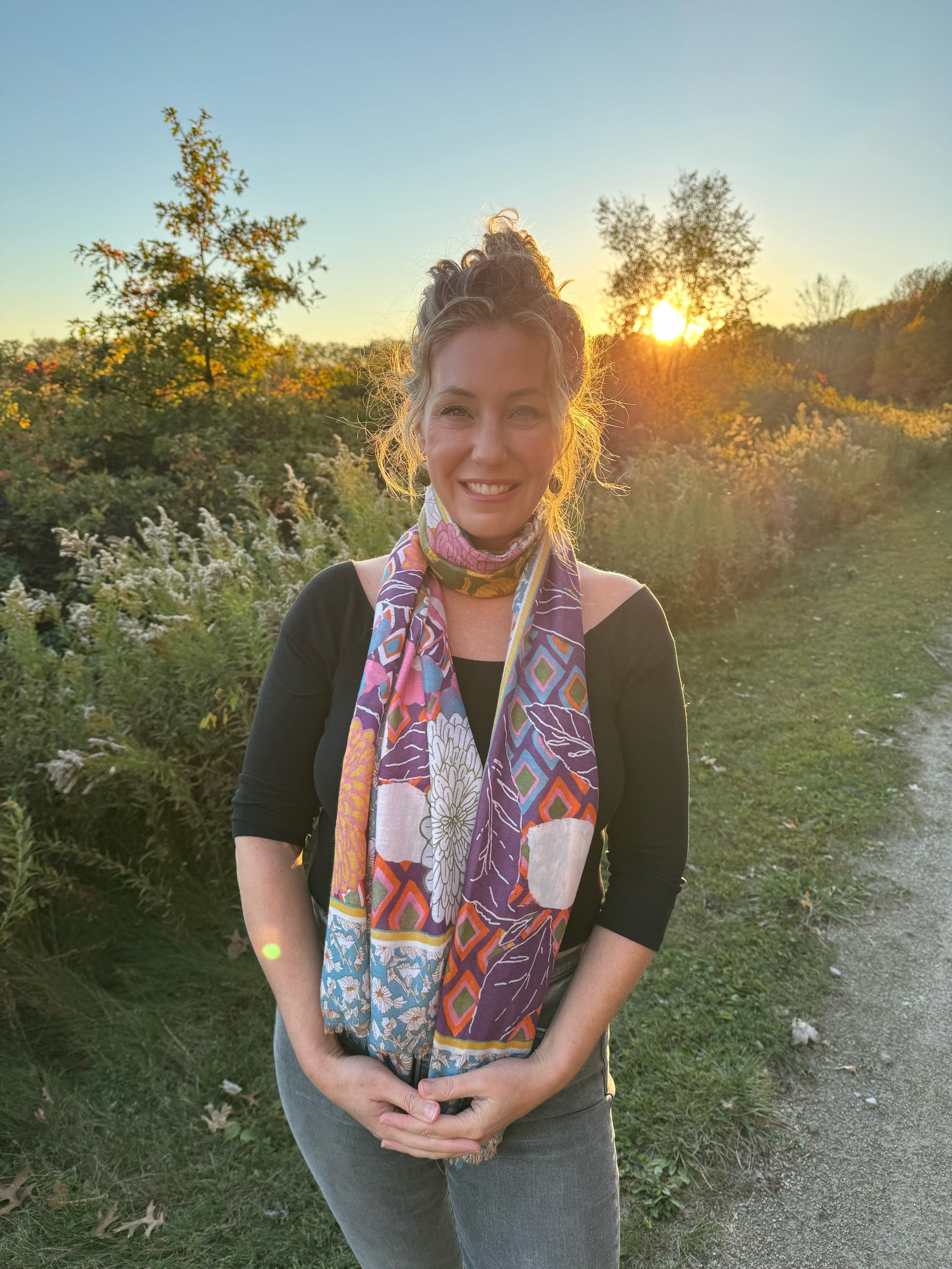 A woman with curly hair in a bun is smiling outdoors at sunset, wearing a colorful scarf and a black top, standing on a trail with greenery and trees in the background.