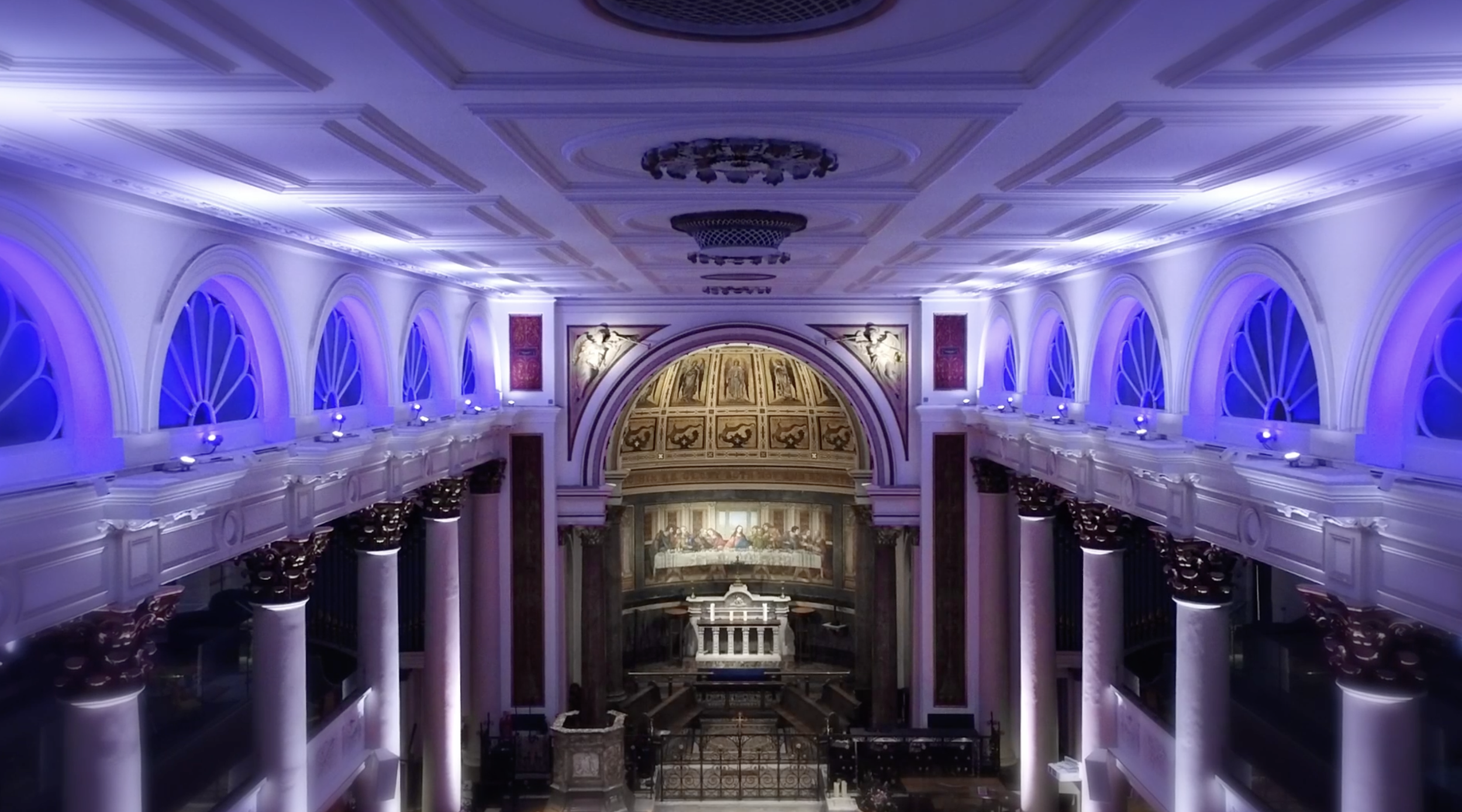 Interior of a grand church with tall columns, arched windows illuminated with purple lighting, and an altar with a painting of the Last Supper by Joe Beynon Media