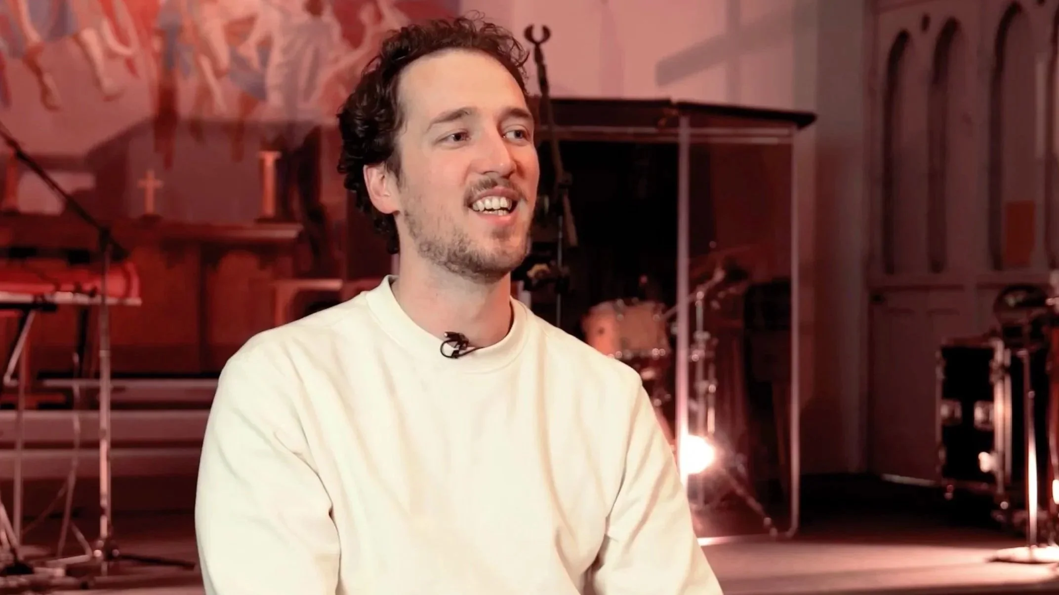 A man with a beard, wearing an off-white sweatshirt, talking head framing, in a church, pinky red backdrop. Joe Beynon Media