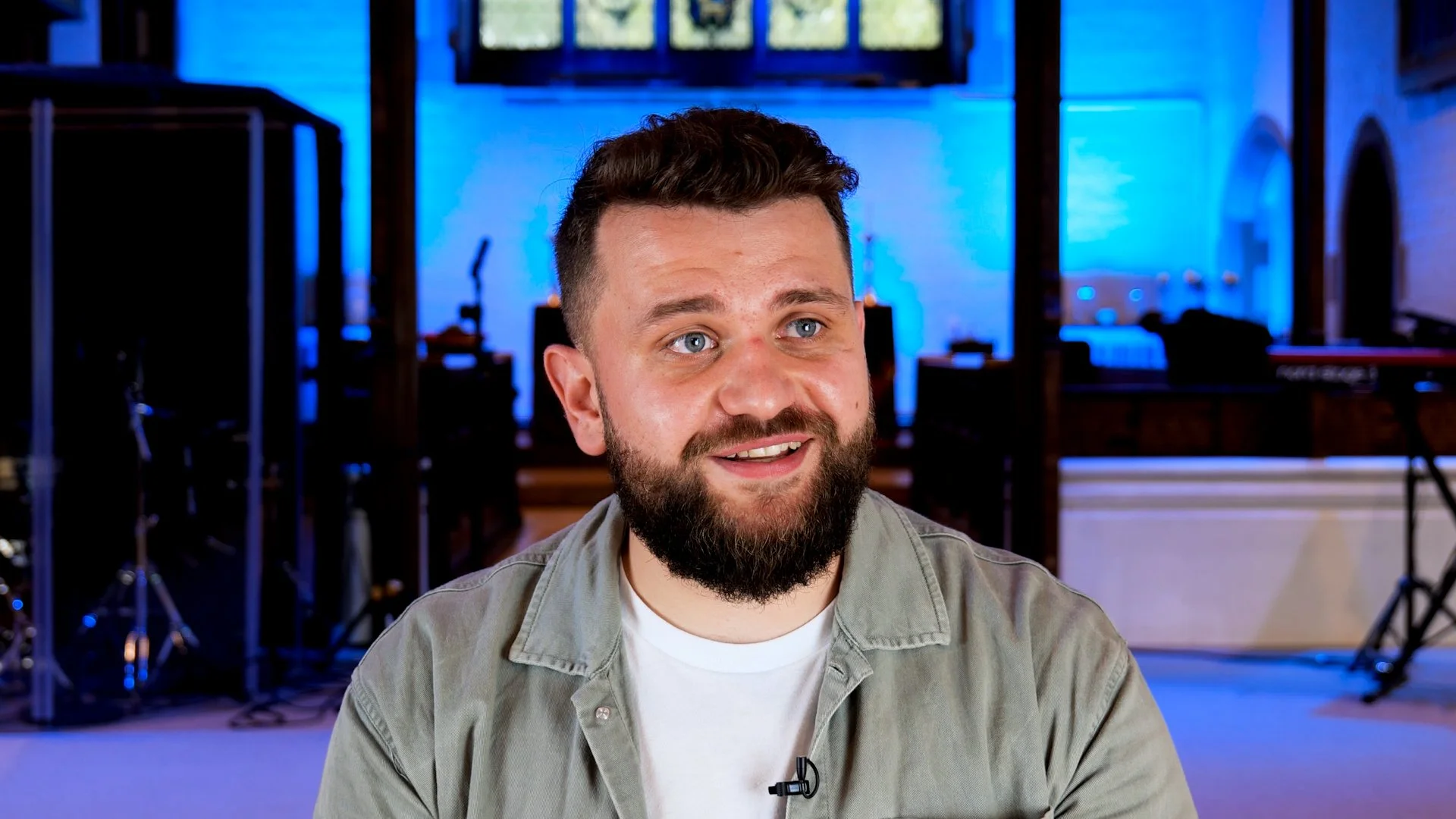 A smiling man with a beard sitting in a church, blue lighting backdrop. Musical instruments and equipment are visible in the background by Joe Beynon Media