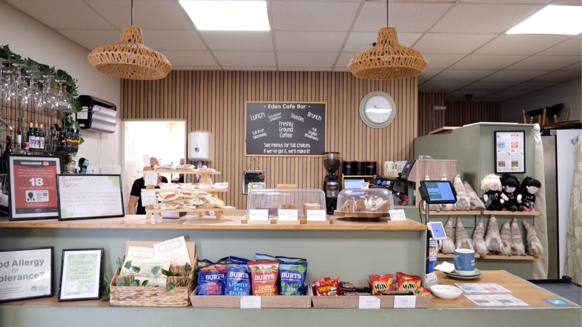 Interior of a green and brown cafe with a counter displaying crisps and baked goods by Joe Beynon Media