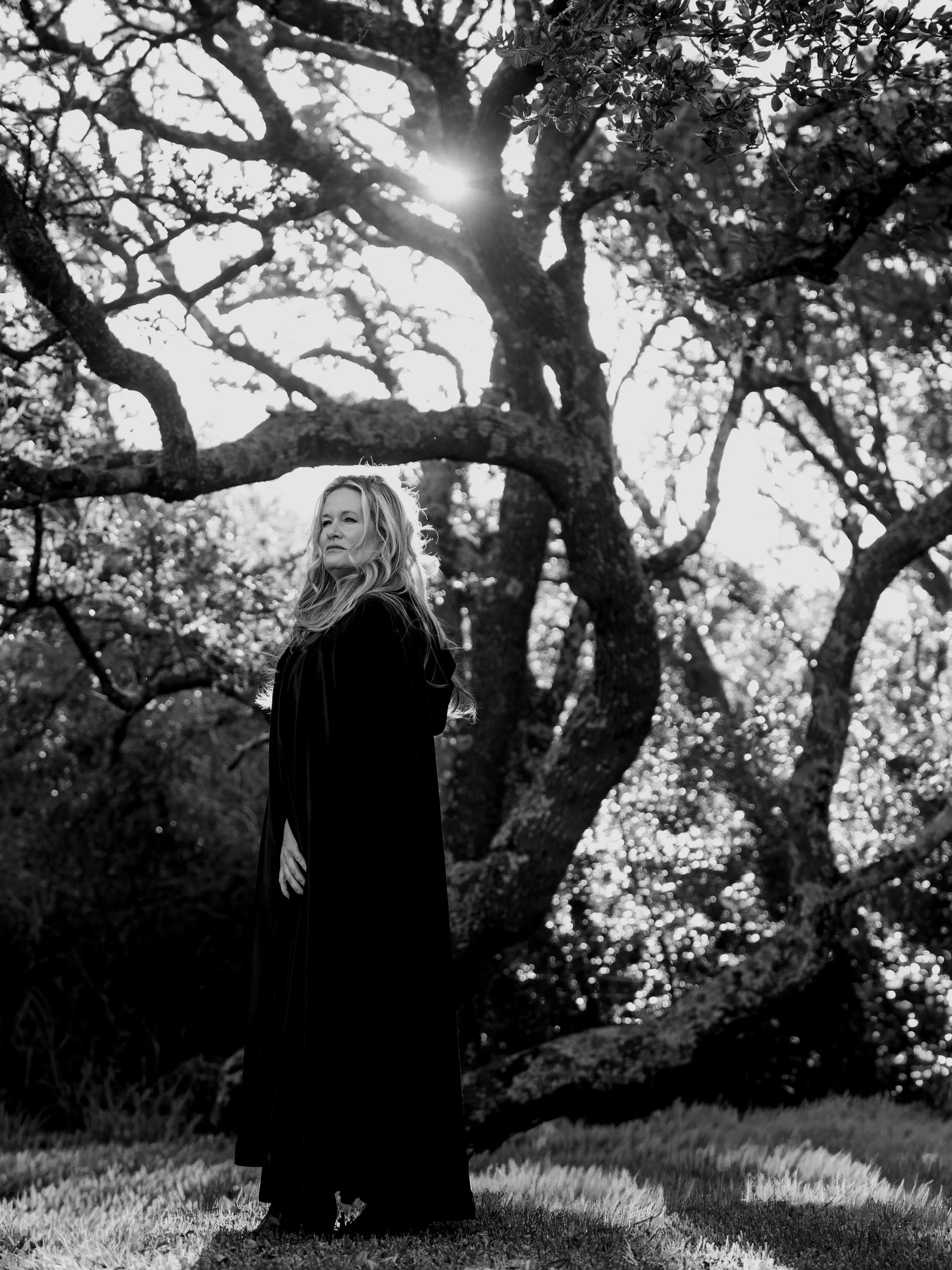A woman with long hair stands outdoors in front of a large, twisted tree with sunlight filtering through its leaves, in a black-and-white photo.