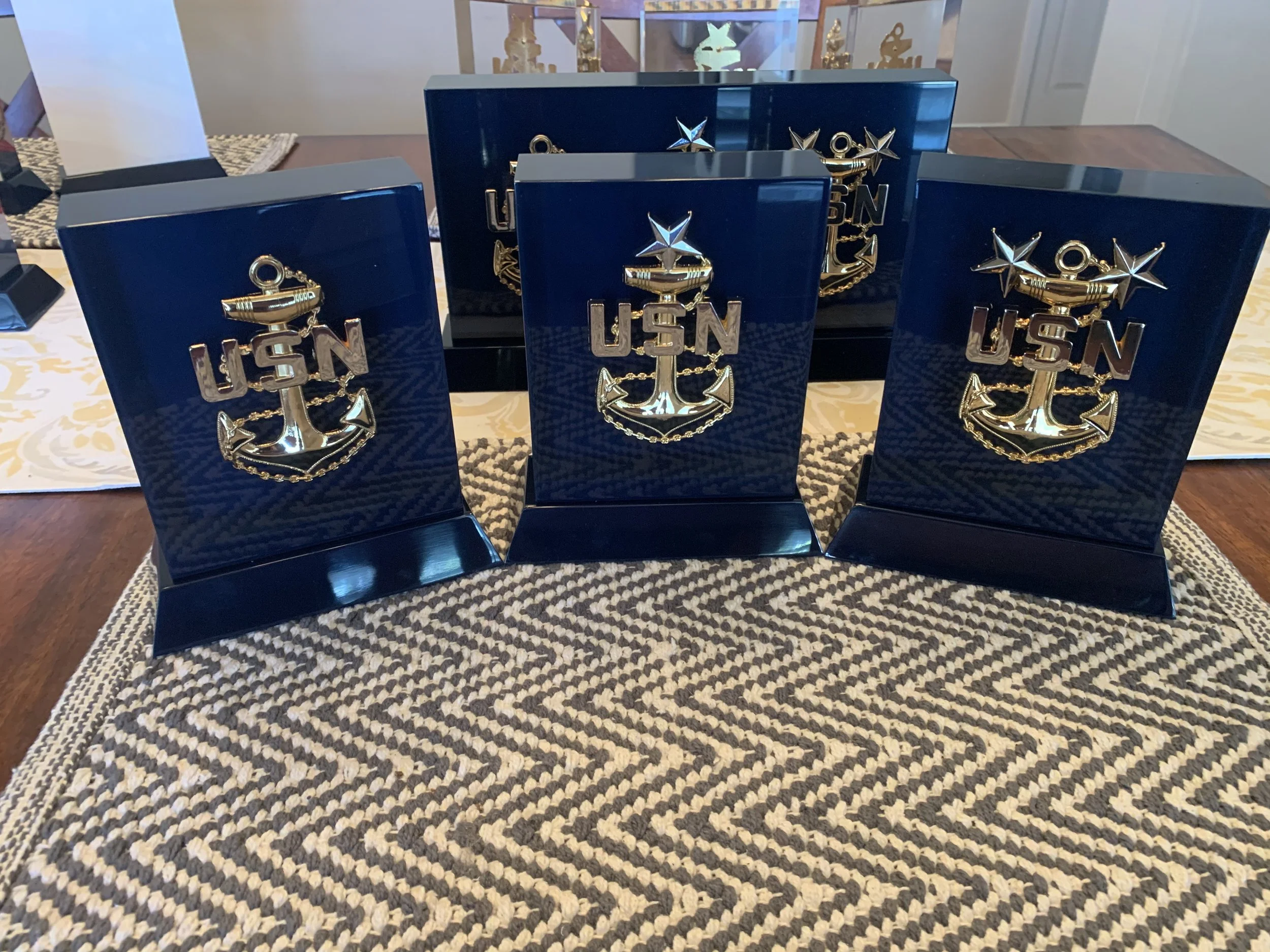 Three navy blue boxes with gold U.S. Navy insignia on a table, with a black display case behind them showing additional navy insignia and awards.