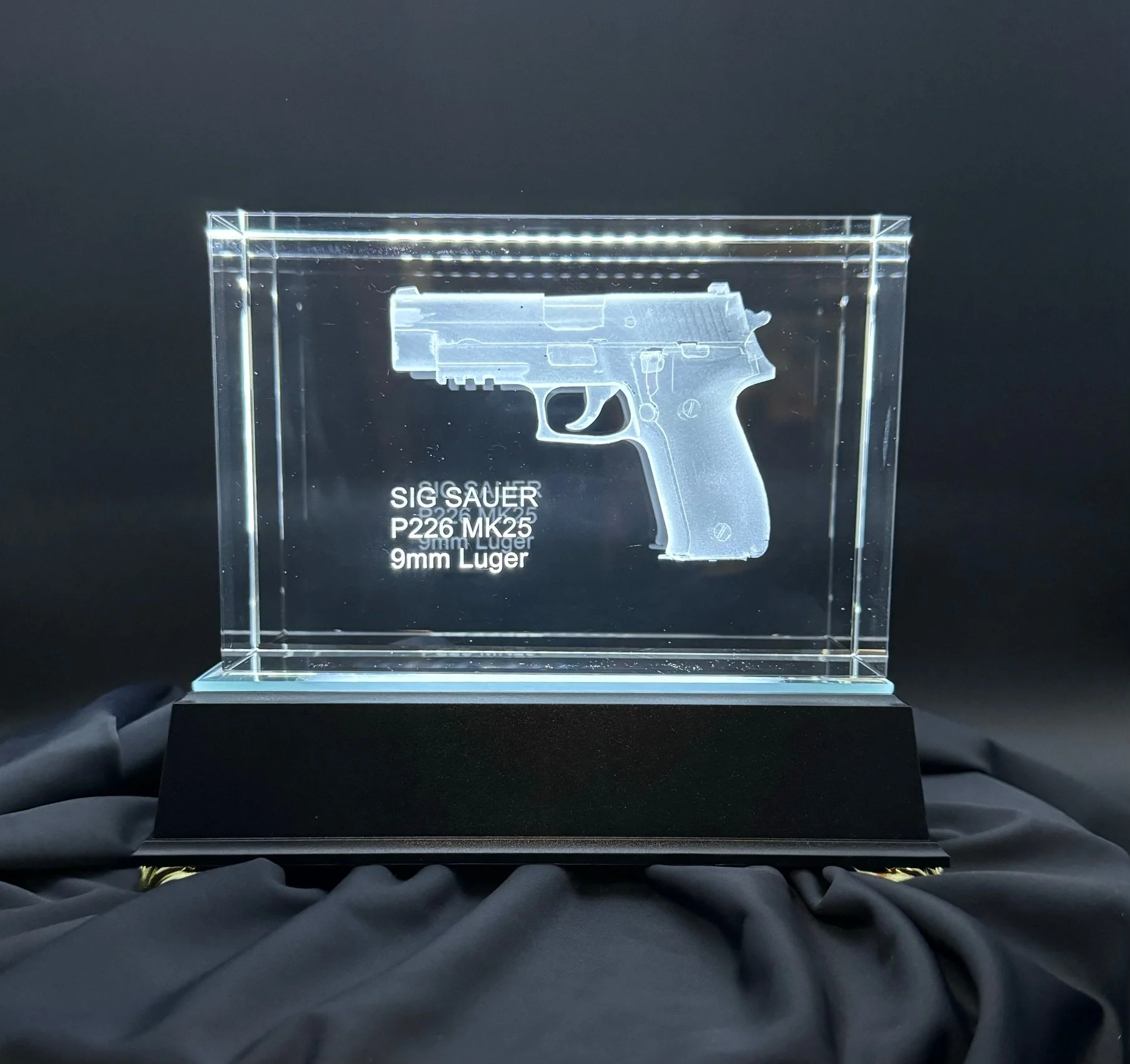 Laser-etched glass display of a SIG Sauer P226 MK25 9mm Luger handgun, displayed on a black base, with black gloves holding it against a dark background.