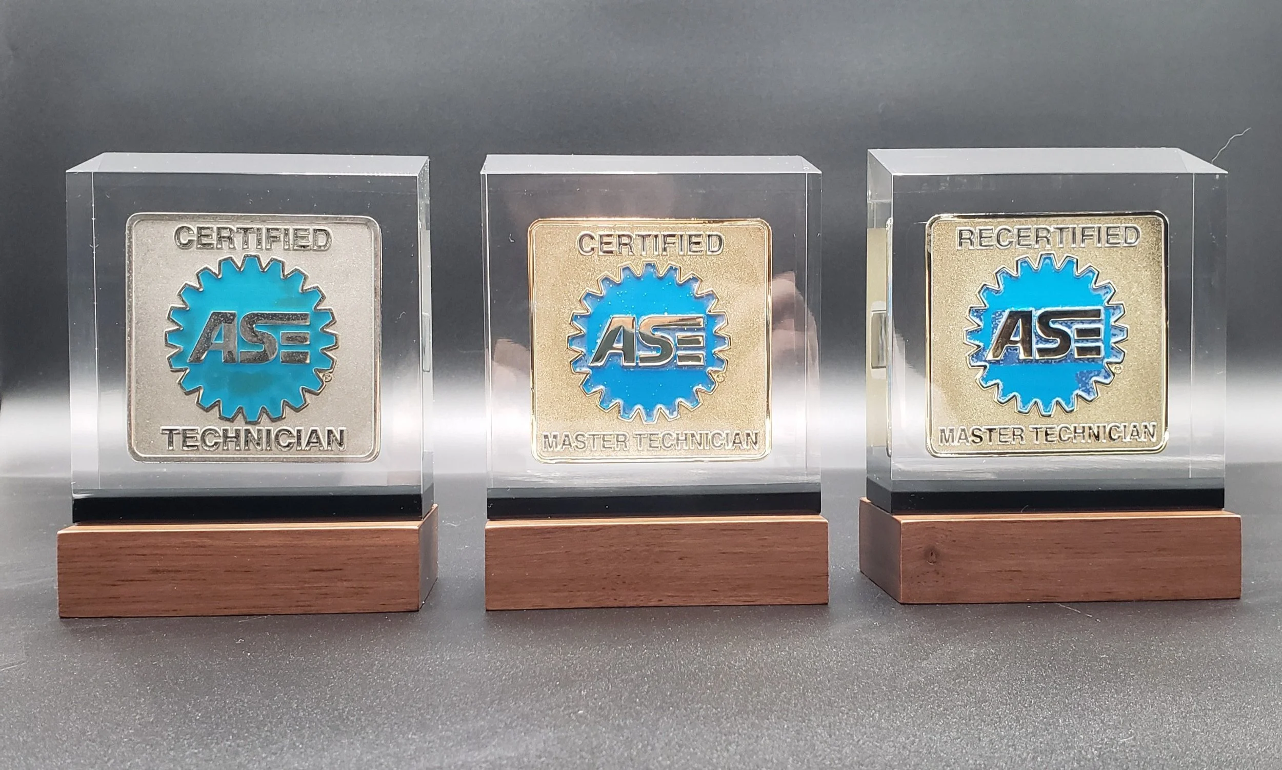 Three glass display cases with ASE certification badges for Technician, Master Technician, and Recertified Master Technician, each mounted on wooden bases.