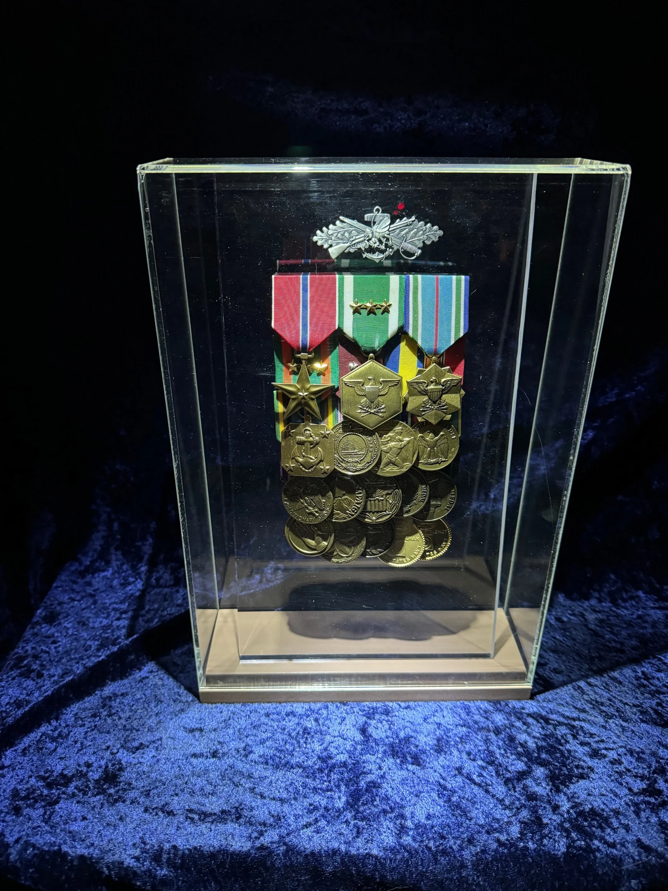 A display case with military medals and badges, illuminated against a dark background with blue lighting on the surface.