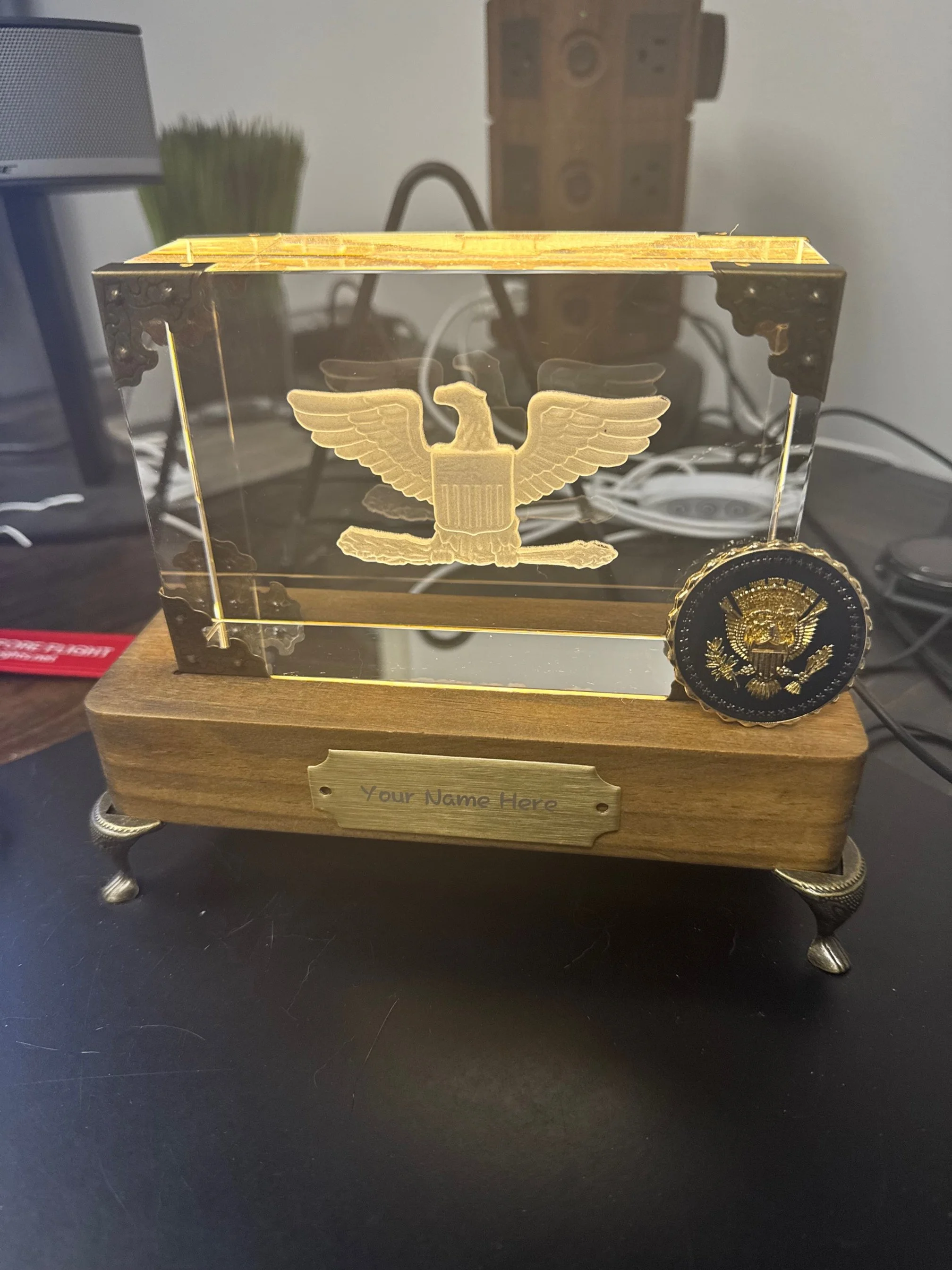 A decorative award with a transparent rectangular case featuring an eagle emblem. The award is mounted on a wooden base with a metal plate that says "Your Name Here". There is a circular seal with a golden eagle emblem attached to the right side of t