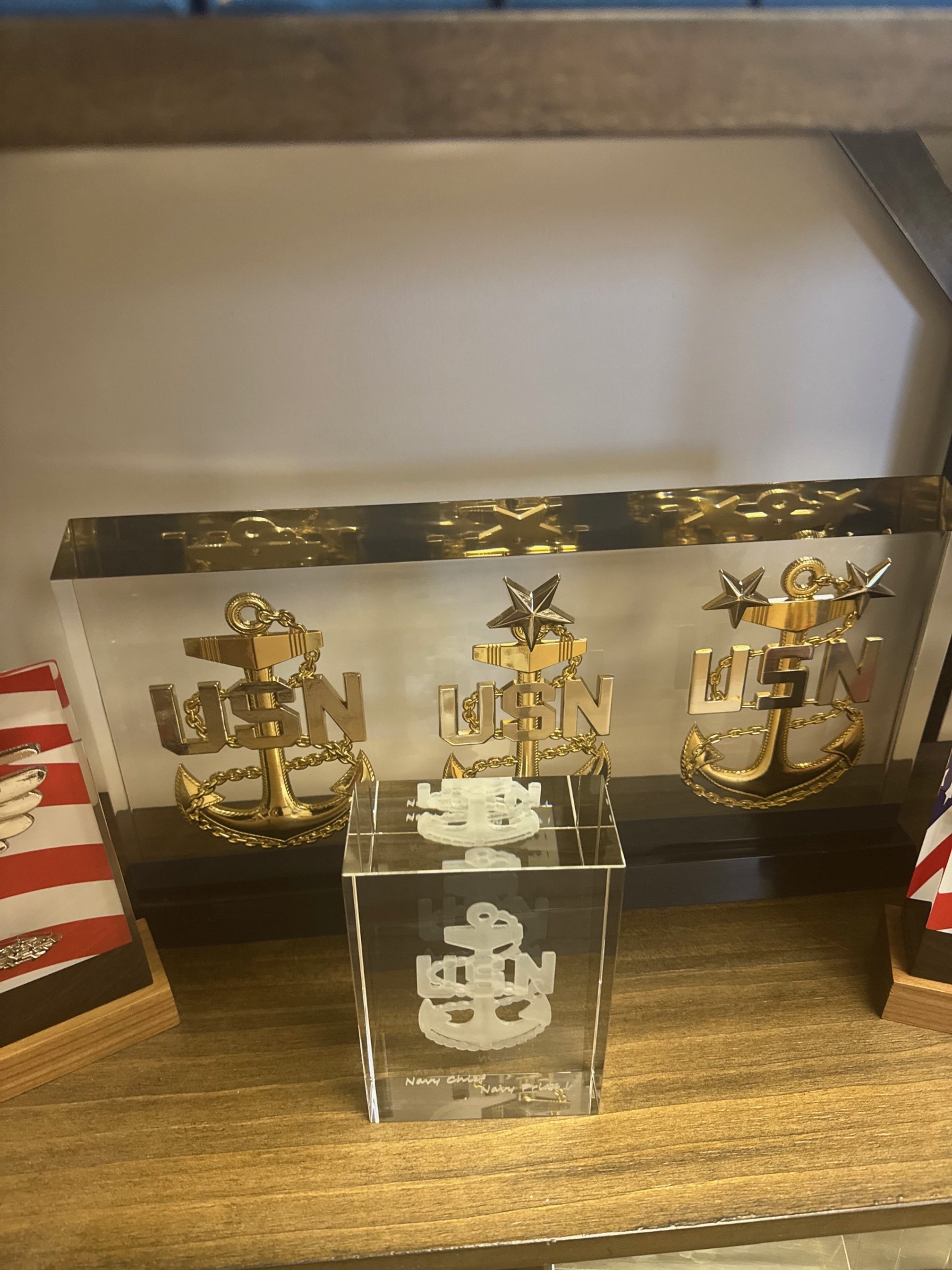Display of three gold U.S. Navy anchor pins with the letters 'USN' and a star, mounted on a clear acrylic stand, on a wooden surface.