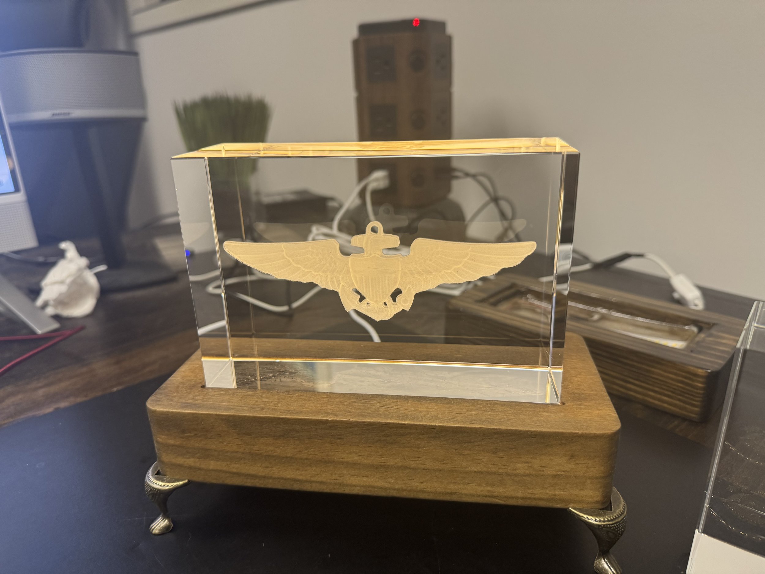 A clear acrylic display case with a wooden base featuring metallic legs, containing a golden eagle emblem with outstretched wings.
