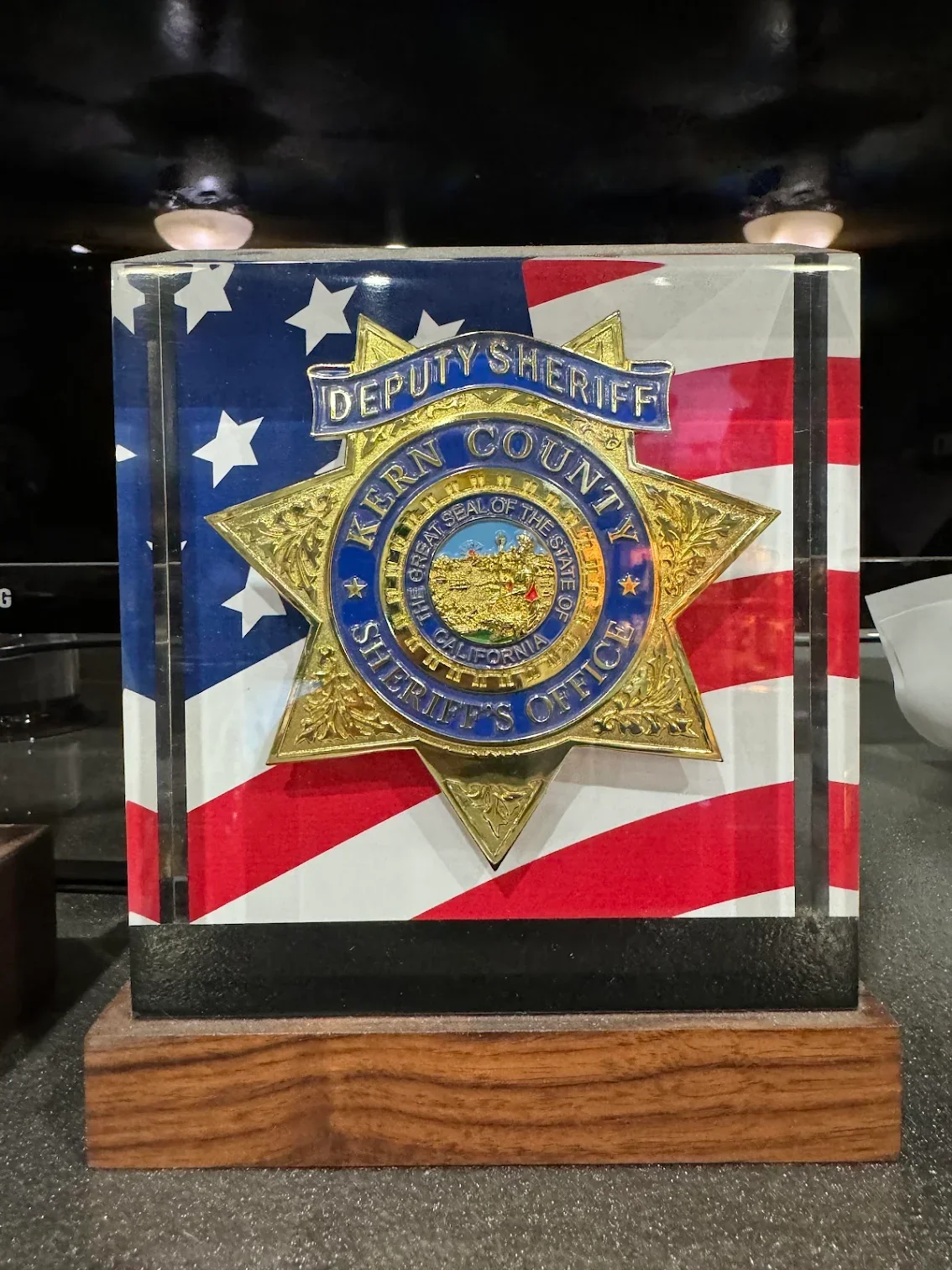 A sheriff's badge with the California state seal, mounted on a display case with an American flag background, labeled 'Lern County Sheriff's Office' and 'Deputy Sheriff'.
