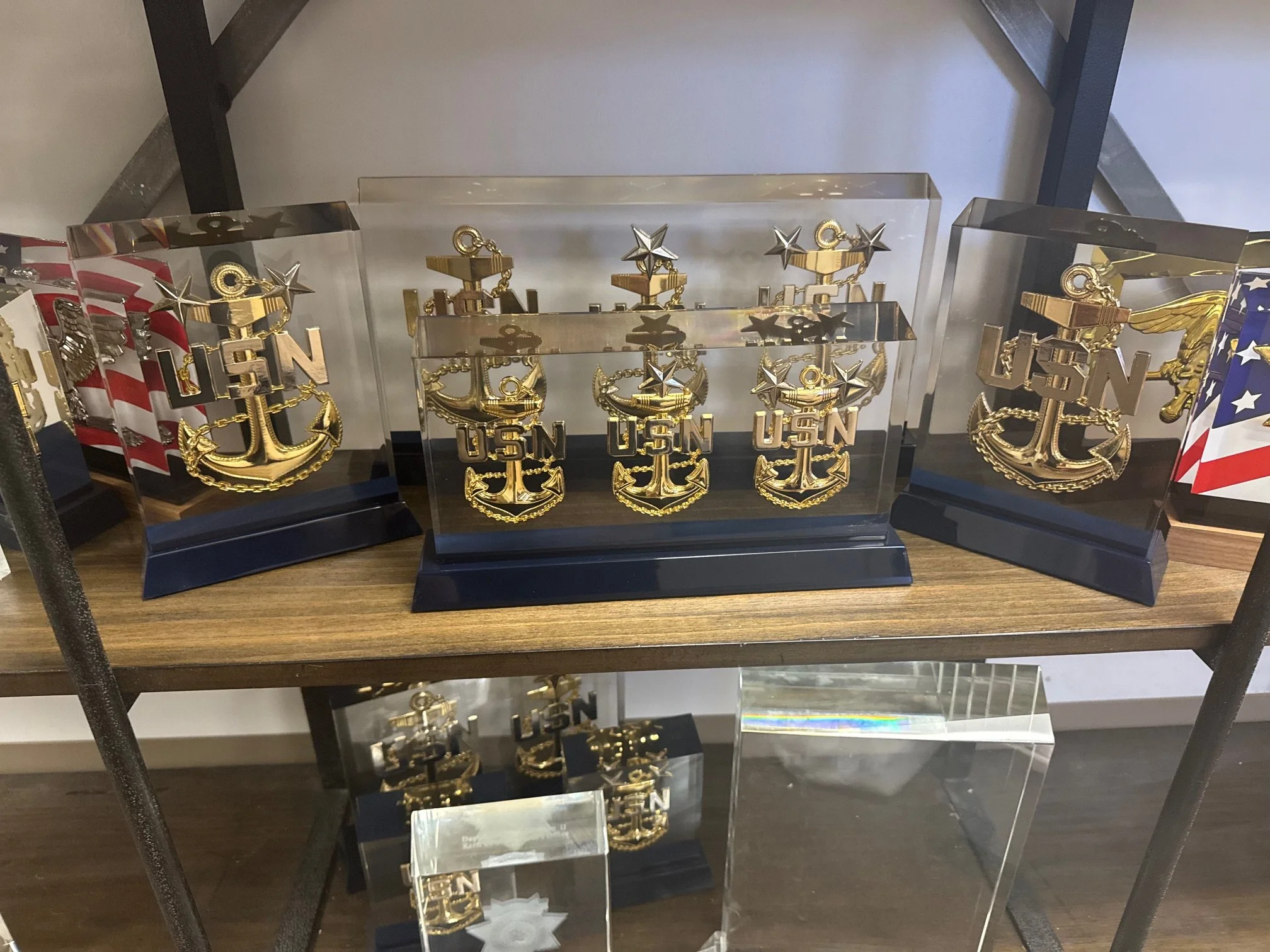 Display of gold-colored USN (United States Navy) anchor emblems with stars, enclosed in clear rectangular cases, arranged on a wooden shelf.