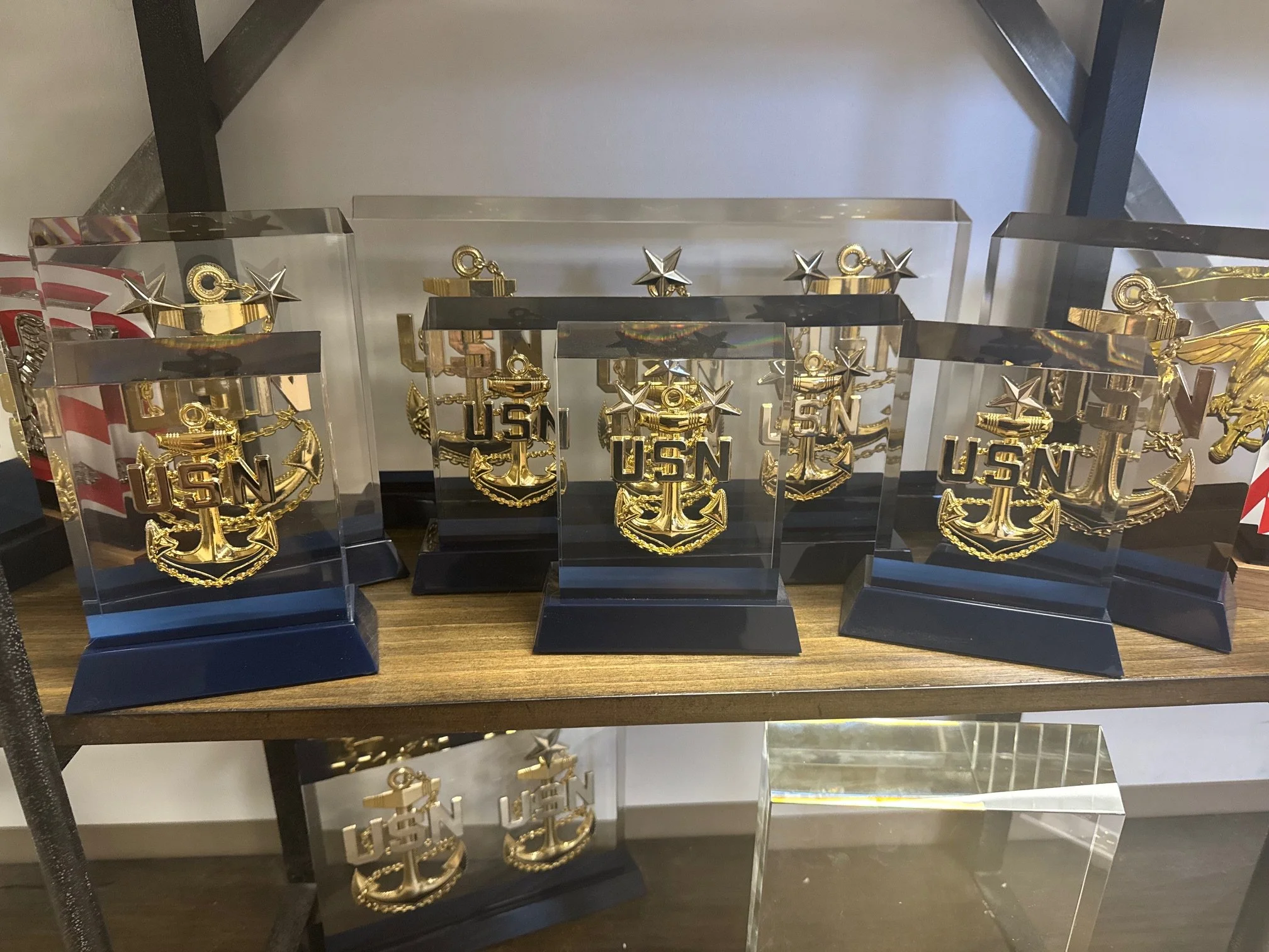 Multiple clear acrylic awards with gold USN (United States Navy) anchors on a wooden shelf.