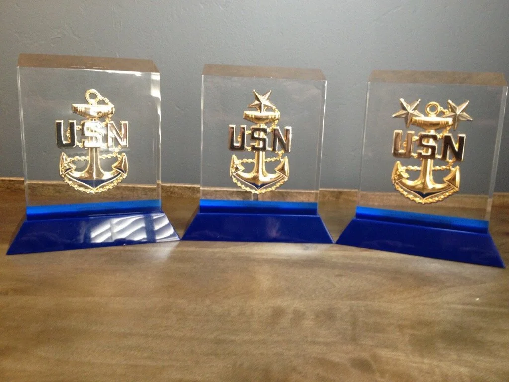 Three gold-colored U.S. Navy anchor emblems with "USN" lettering, mounted on acrylic plaques with blue bases, placed on a wooden surface.