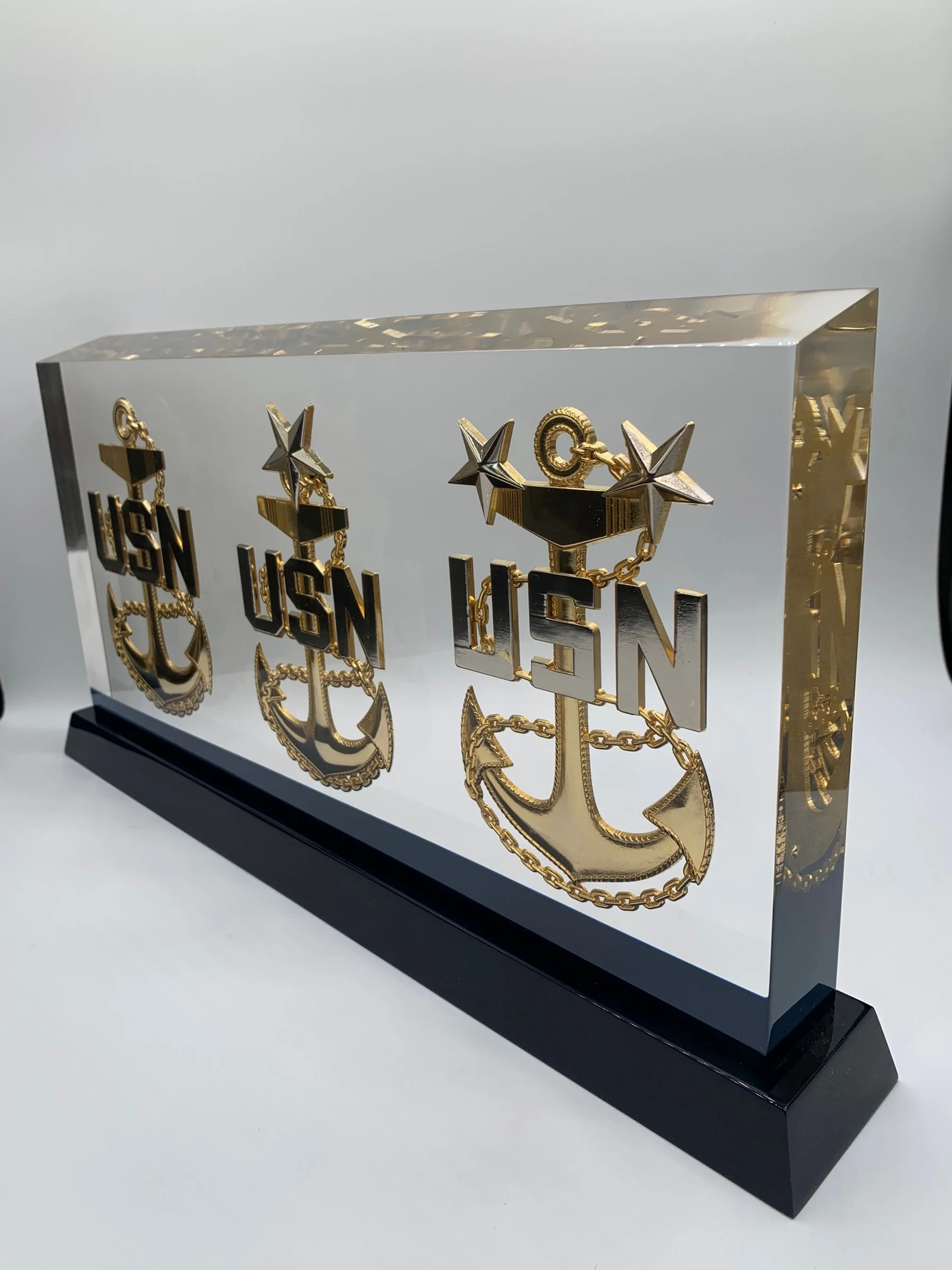Decorative display of three gold and black US Navy emblems with an anchor, stars, and chains on a clear acrylic background with a black base.