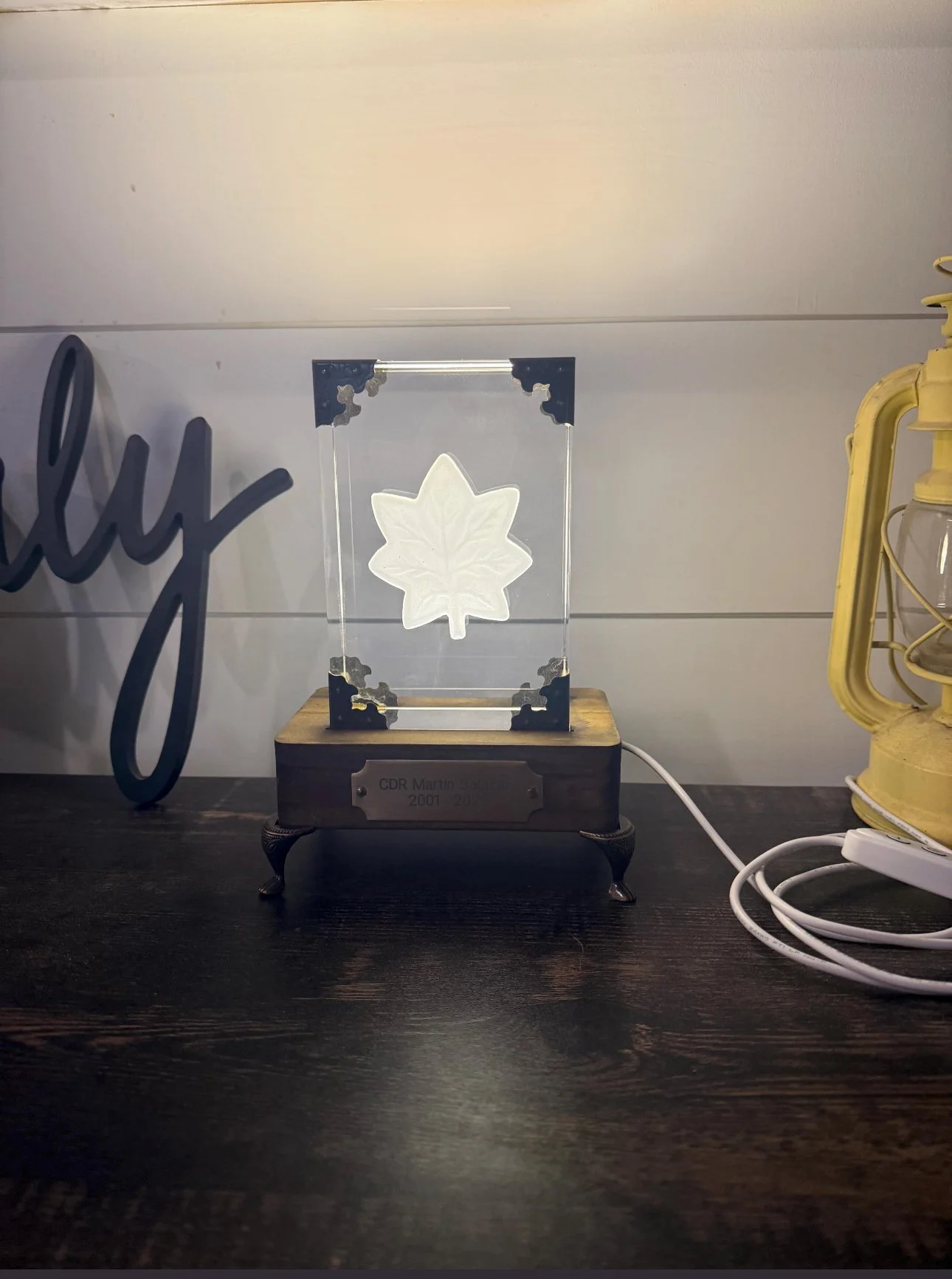 Decorative illuminated acrylic block with a carved maple leaf design, mounted on a wooden base with a metal plaque, placed on a dark wooden surface beside a yellow lantern and coiled white electrical cord.