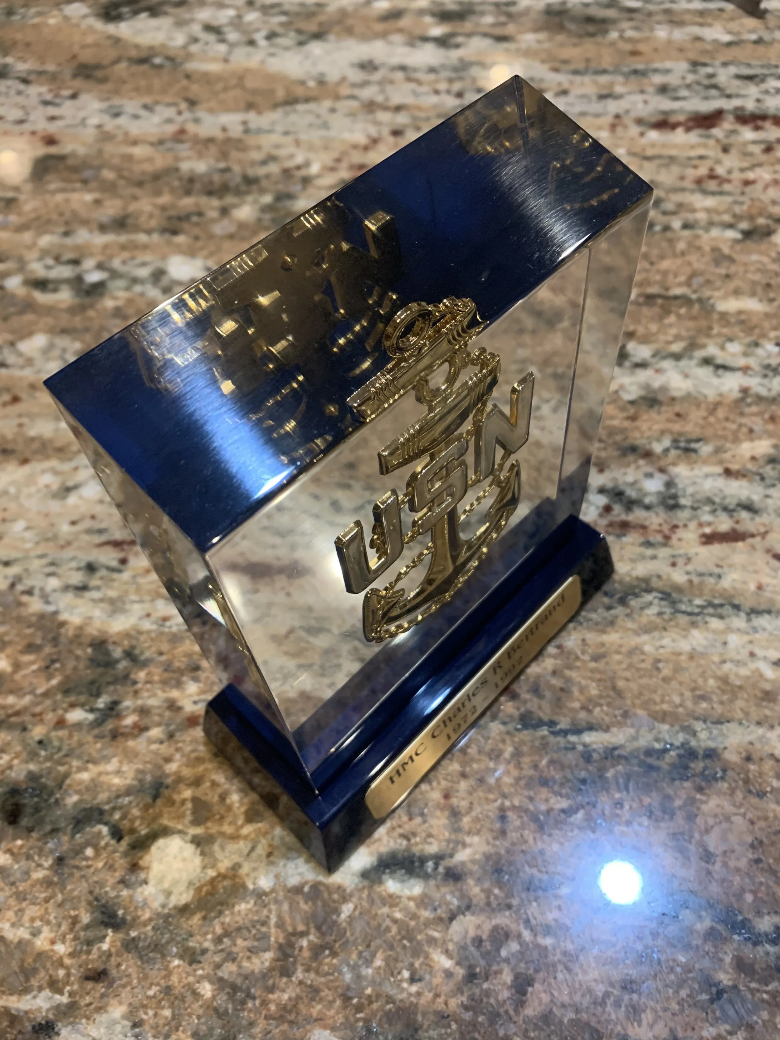 A clear acrylic award with a dark blue base, featuring a gold emblem and plaque. The plaque reads 'HMC CHARLES R. BERTRAND 1929-2018' on a marble countertop.