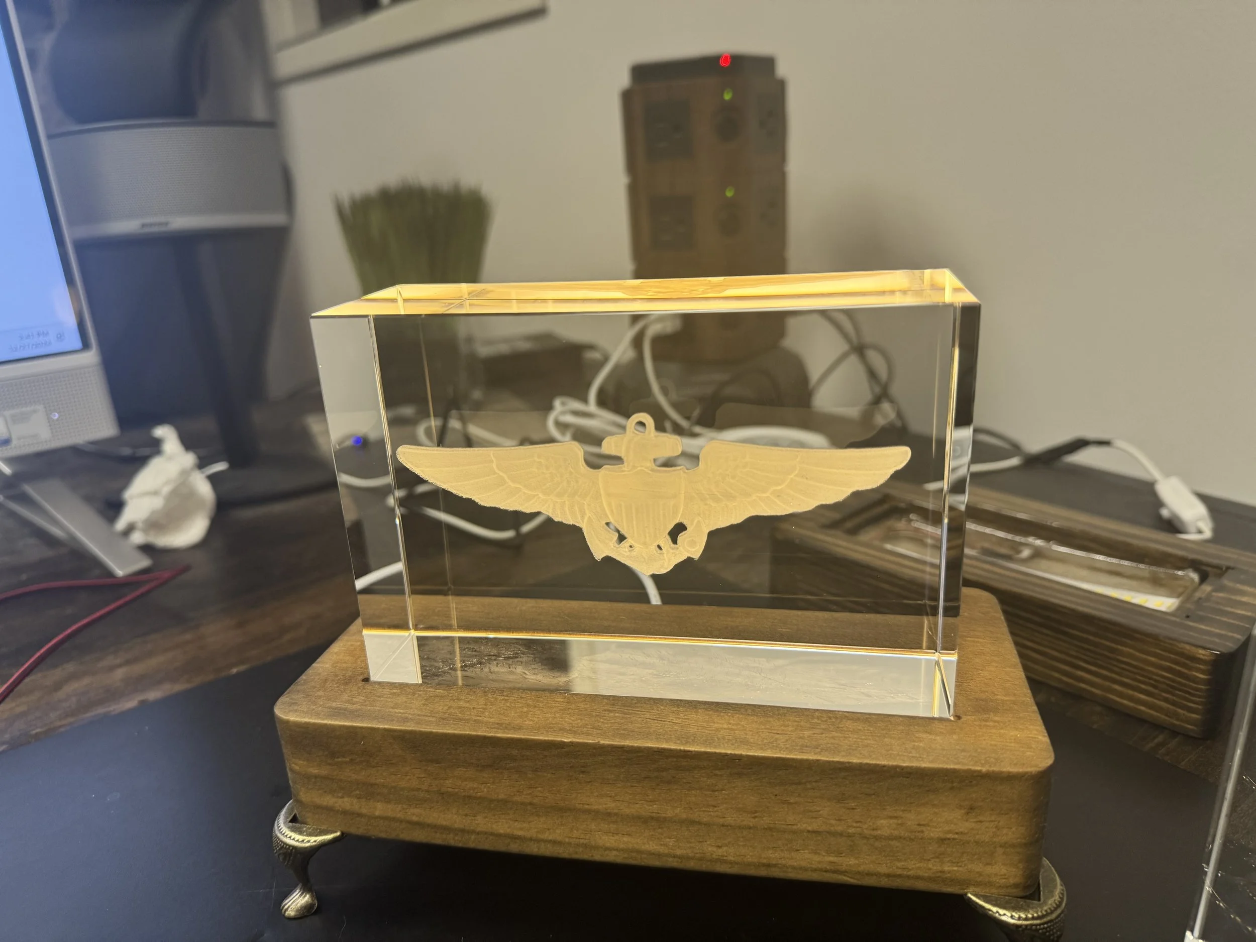 A golden eagle insignia encased in a clear rectangular acrylic display, mounted on a wooden base with metallic decorative feet, placed on a dark surface.