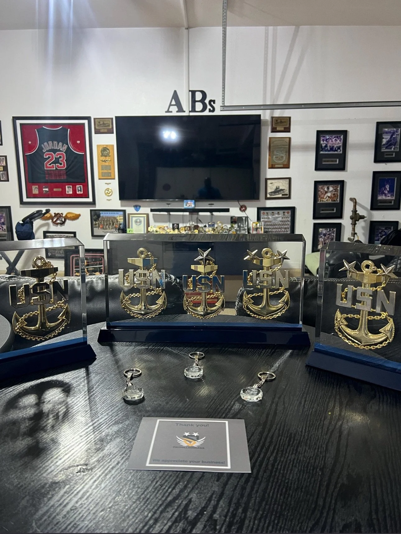 Four glass plaques with gold anchors and 'USN' initials inside plastic cases on a table. Four keychains with clear crystals on the table. Framed sports memorabilia, including a Michael Jordan jersey, and a large flat-screen TV on the wall in the background.