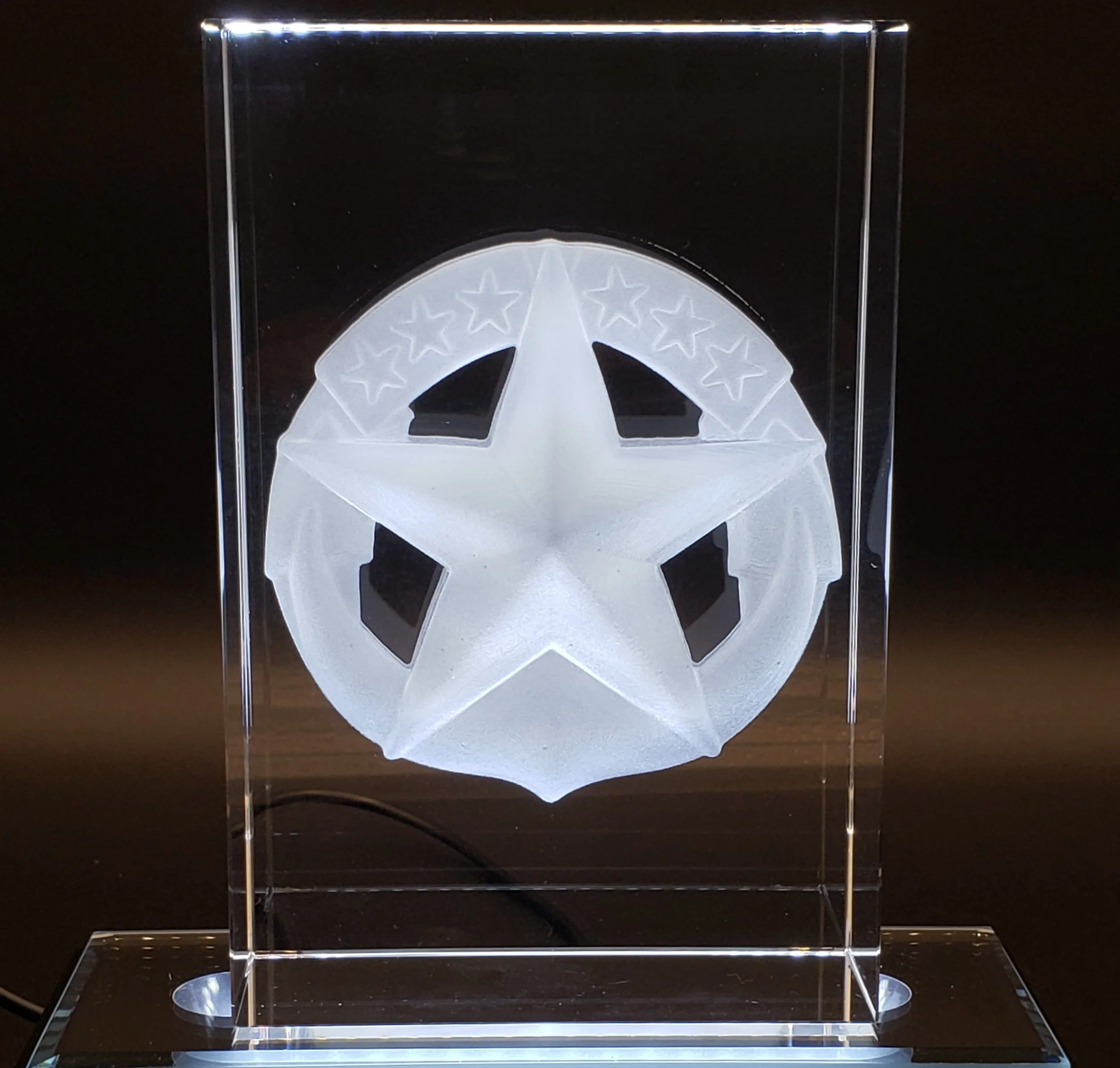 A clear acrylic block with a white star and circular pattern inside, illuminated from below, mounted on a glass base.