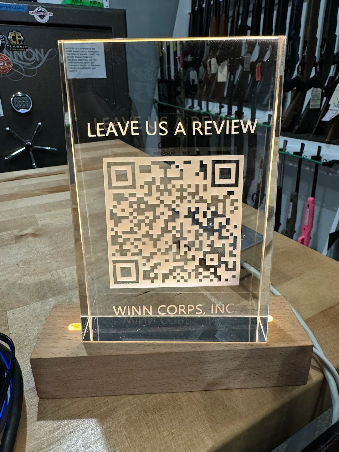 A clear acrylic sign with a gold border on a wooden base, encouraging customers to leave a review by scanning a QR code. The sign reads, 'LEAVE US A REVIEW,' with the company's name 'WINN CORPS, INC' at the bottom, located on a wooden tabletop in a retail or firearm store.