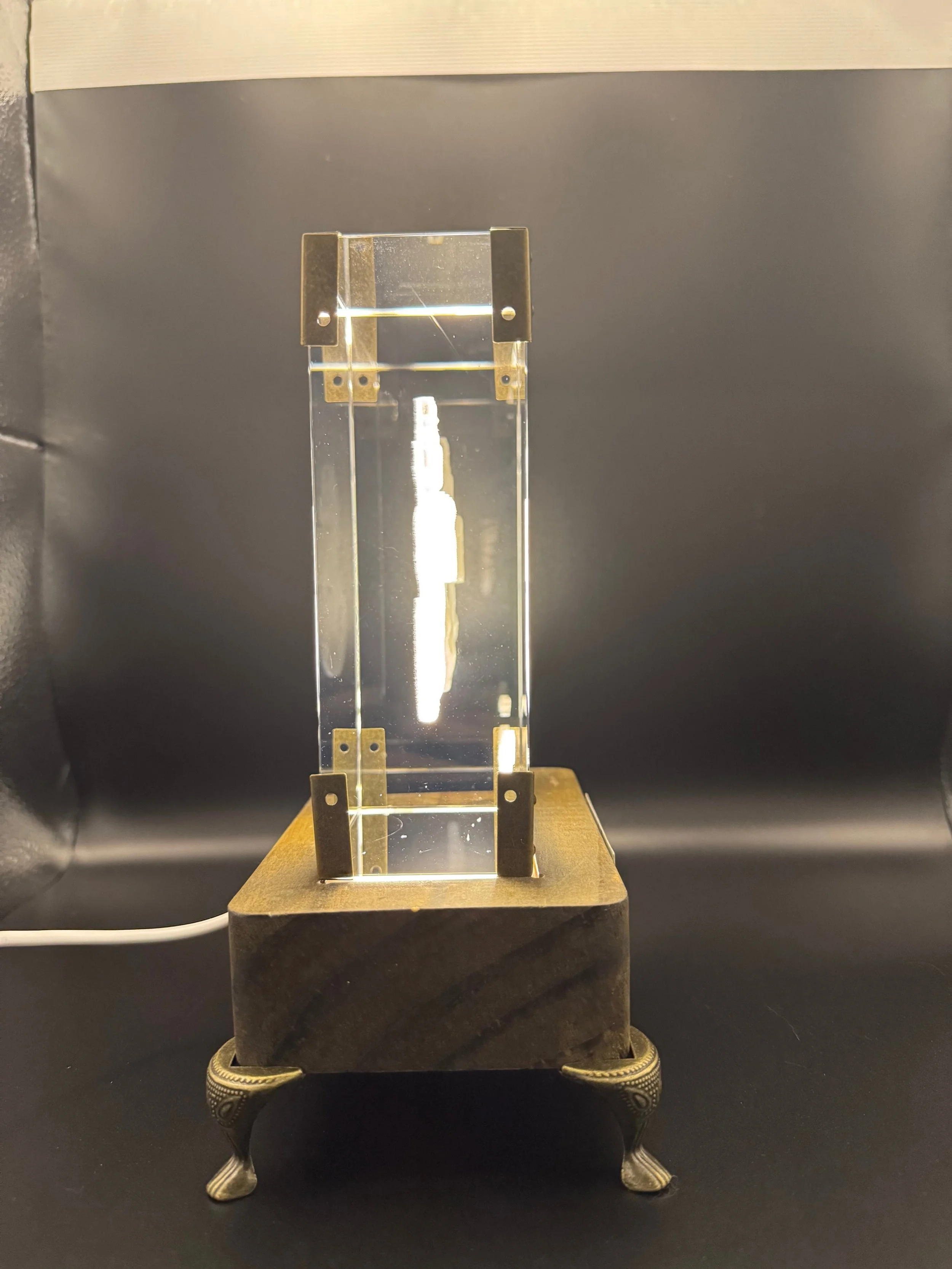 A crystal clear transparent rectangular block with gold-colored metal brackets at the top and bottom, mounted on a wooden stand with ornate legs, against a black backdrop.