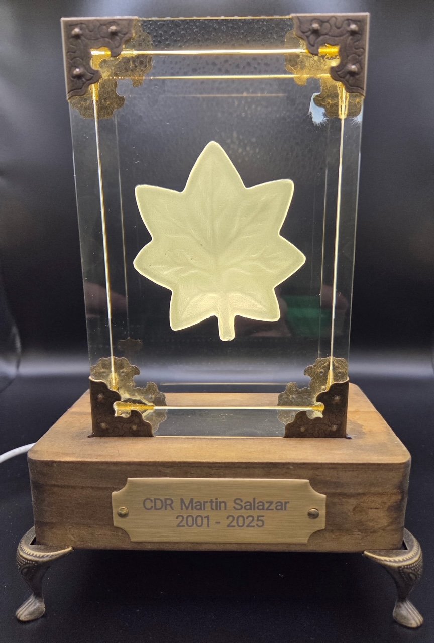 A decorative glass tribute with a maple leaf etched inside, mounted on a wooden base with a brass plaque that reads 'CDR Martin Salazar 2001 - 2025'.