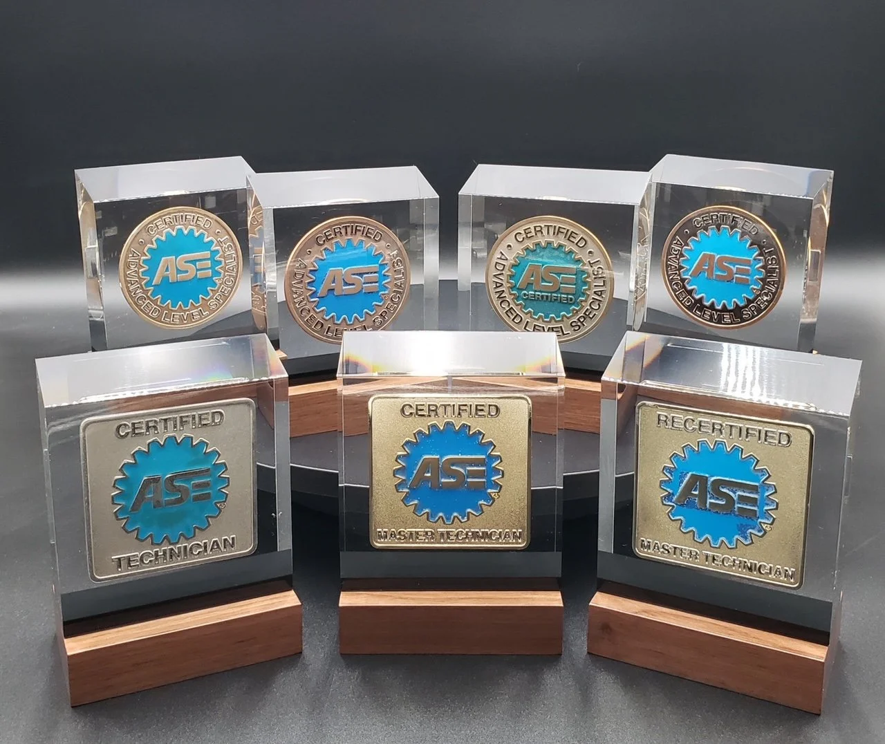 Six BallistiGlass awards with blue and gold ASE logos, displayed on black and wooden bases, with some encased in clear square or rectangular blocks.