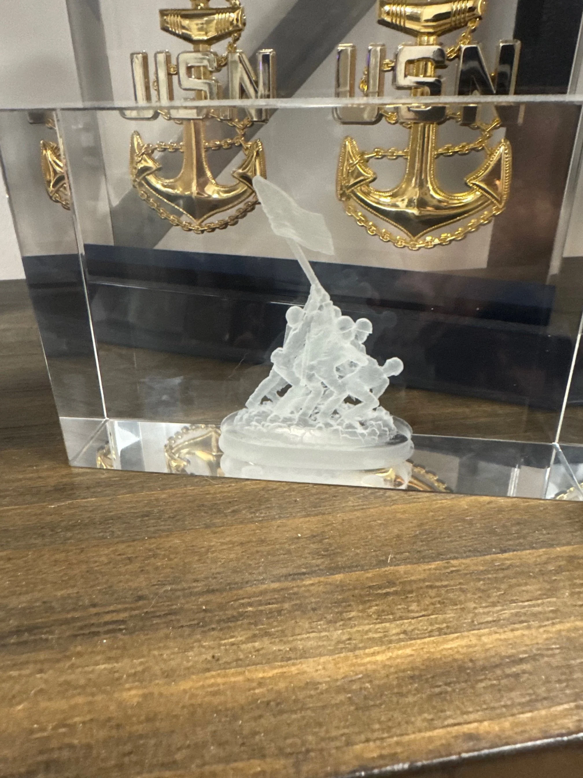 A crystal engraving of soldiers raising a flag, set on a wooden surface with golden anchor decorations in the background.