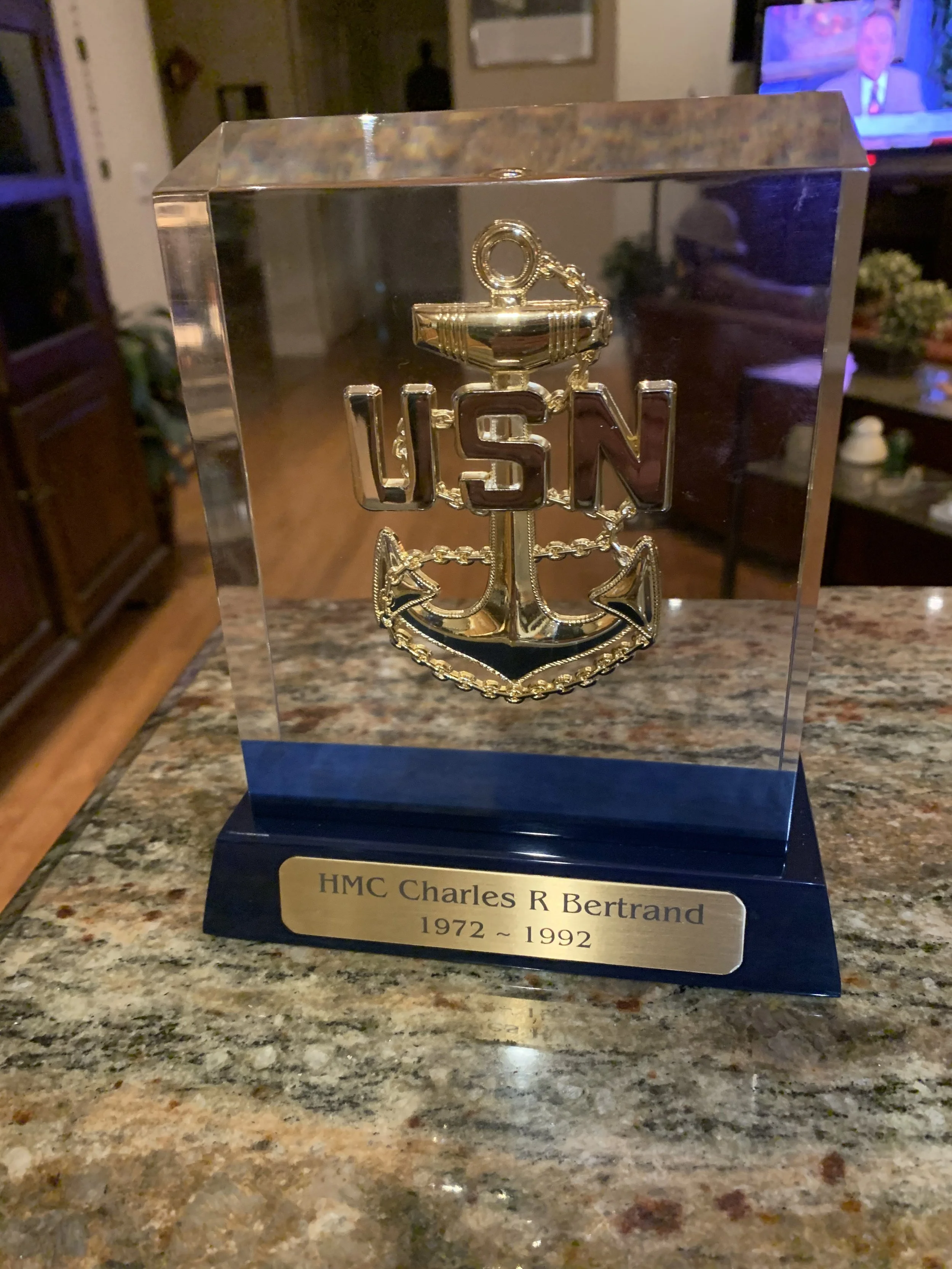 A glass display case with a gold and black anchor badge with the letters USN on it. The case has a plaque that reads 'HMC Charles R Bertrand 1972 - 1992' and is placed on a granite countertop.