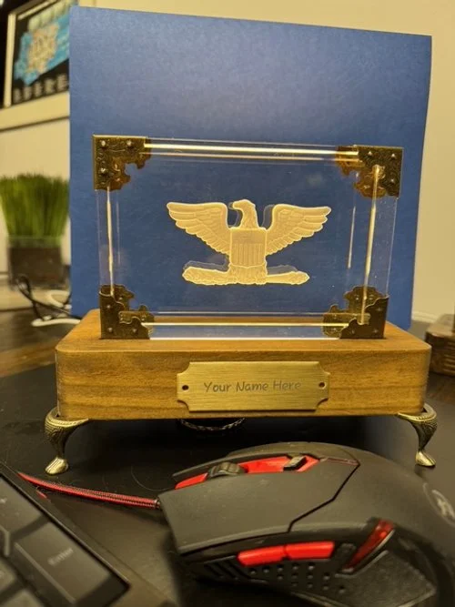 A clear glass display case with a golden eagle emblem inside, mounted on a wooden base with a plaque that says 'Your Name Here'.