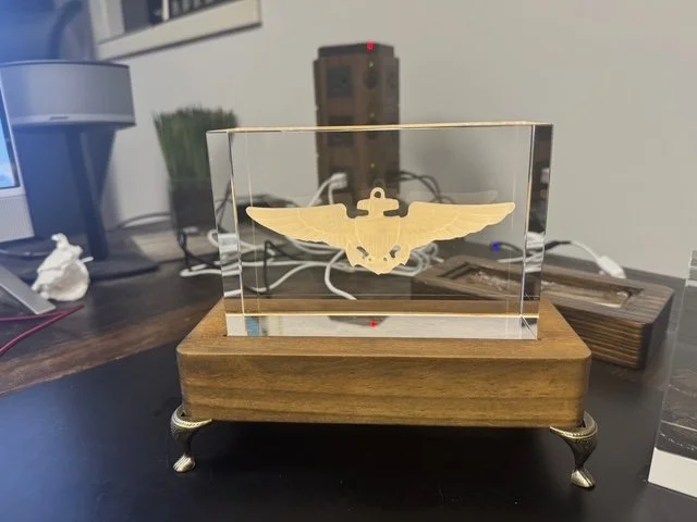 A glass display case with a gold emblem of a bird with outstretched wings and a gear in the center, mounted on a wooden base with metal legs, placed on a dark wooden table.
