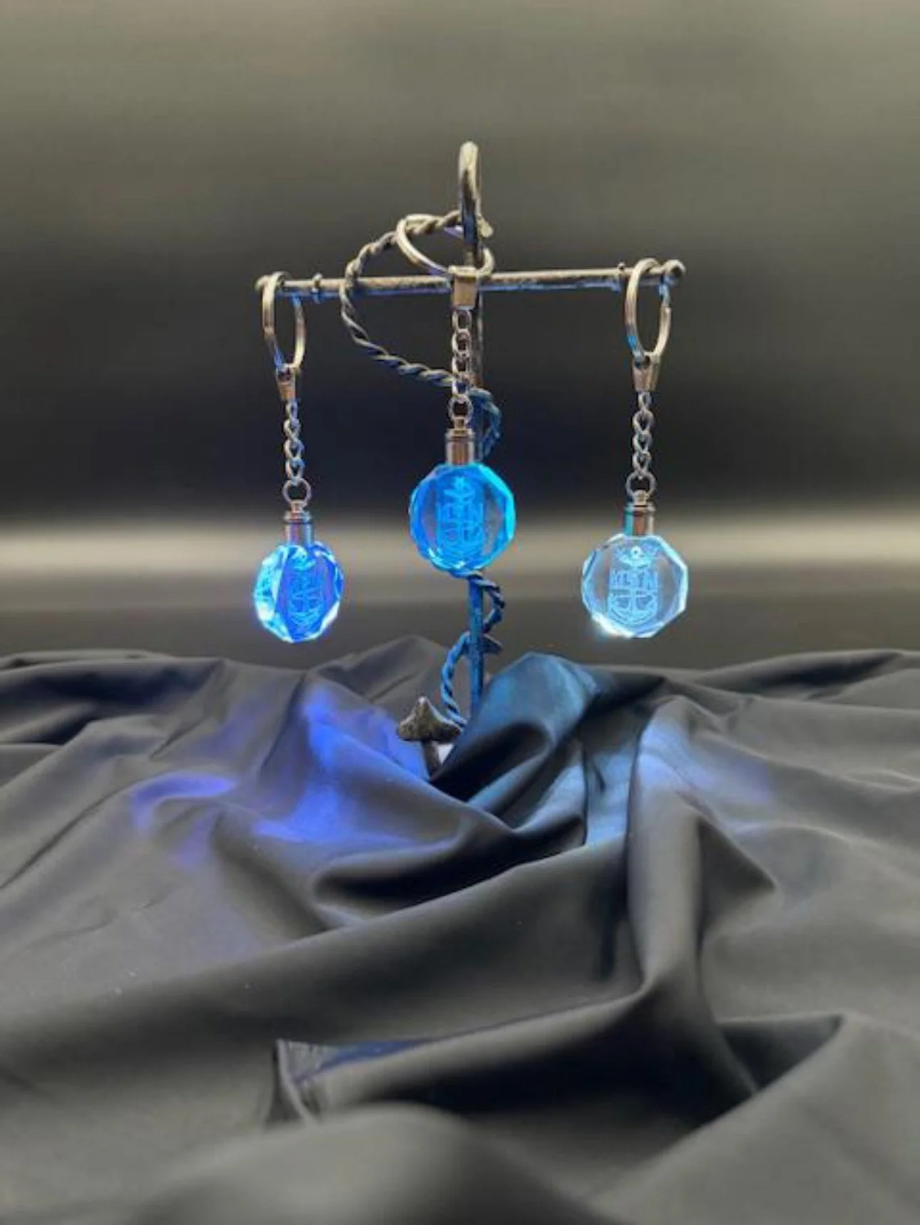 A metallic jewelry display stand holding three blue and clear crystal pendants with chains, set against a dark background and draped in black cloth.