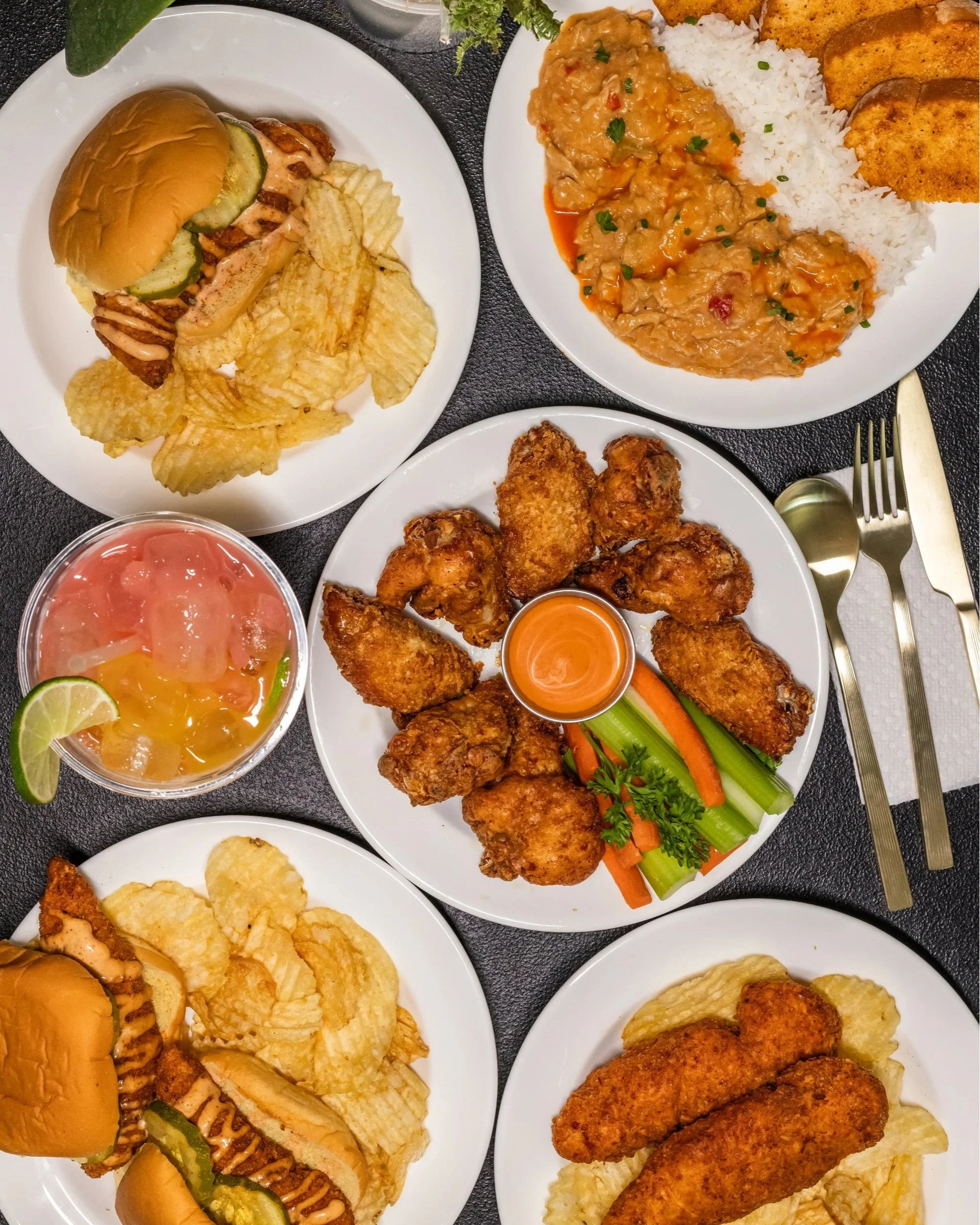 A variety of American dishes on white plates including sandwiches with pickles and chips, fried chicken wings with dipping sauce, fried fish fillets with potato chips, rice with curry and fried chicken breasts, and a bowl of fruit punch with lime slices.