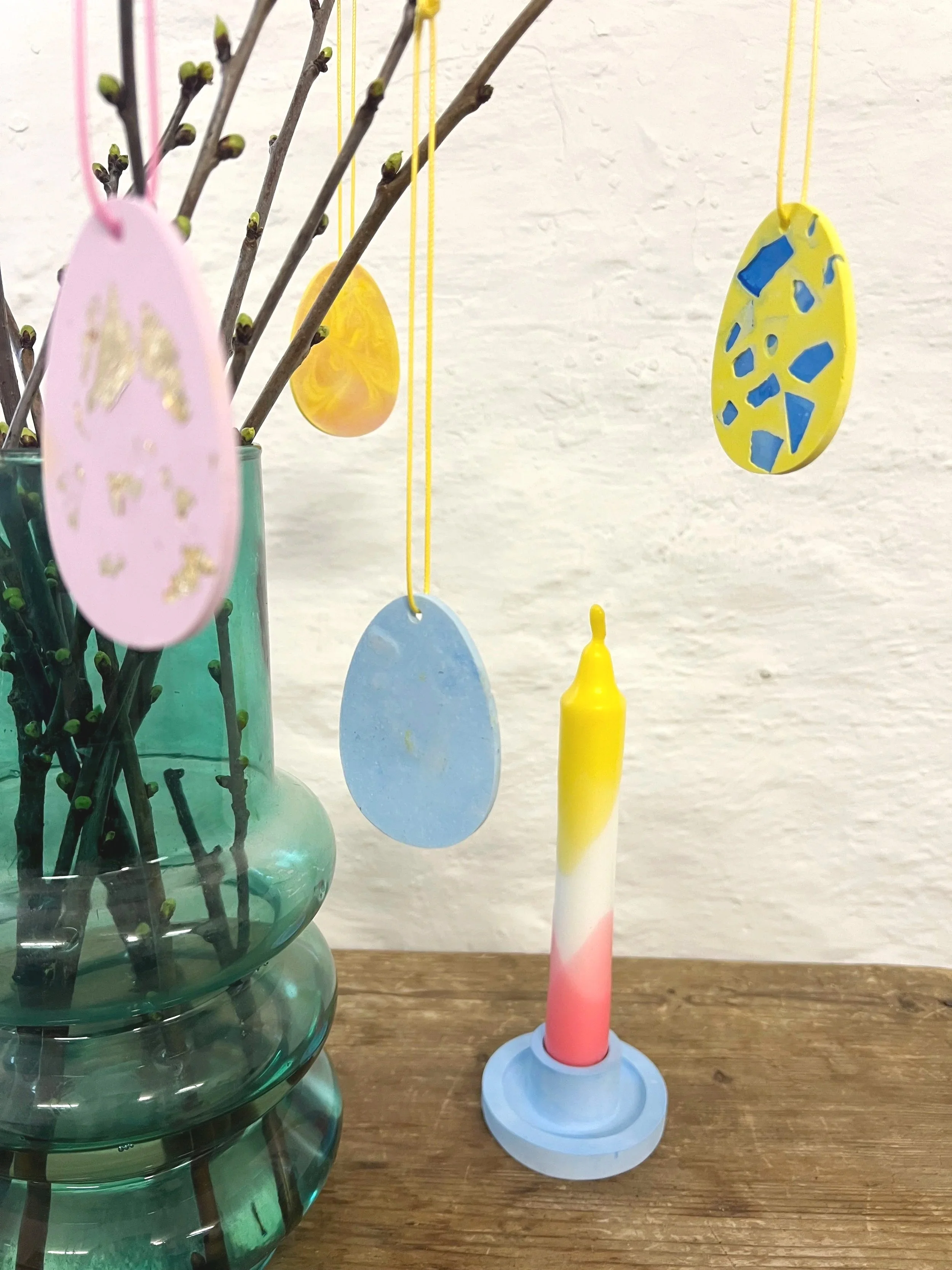 Ostereier aus Keramik mit goldener und blauer Verzierung hängen an Zweigen in einer Vase. Auf einem Holz Tisch steht eine gemusterte Kerze in einem Kerzenständer.