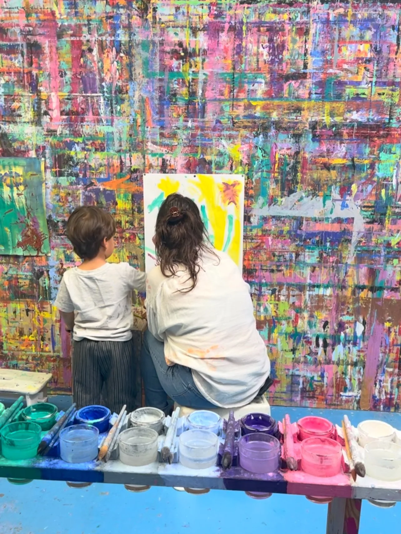 Eine Frau und ein Kind beim Malen in einer kunterbunten Kunstgalerie, vor bunter Wand mit mehreren Gemälden, mit Farbtöpfen und Pinseln auf einem Tisch davor.