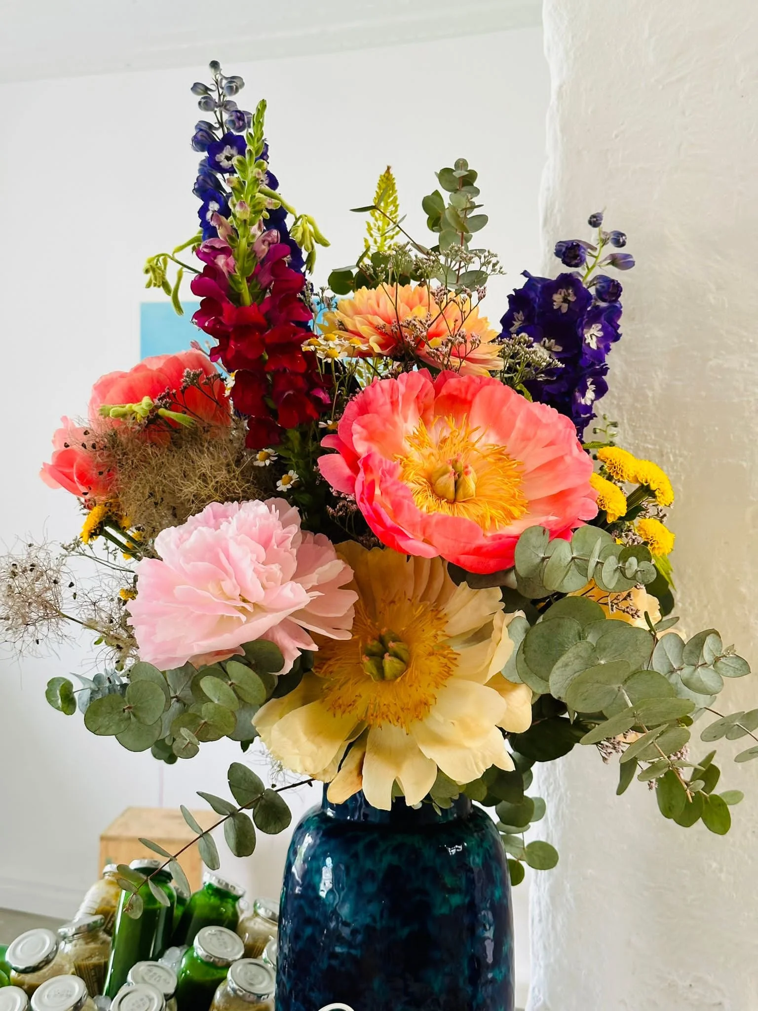 Bunte Blumen in einer blauen Vase, darunter kleinere Gegenstände und Flaschen