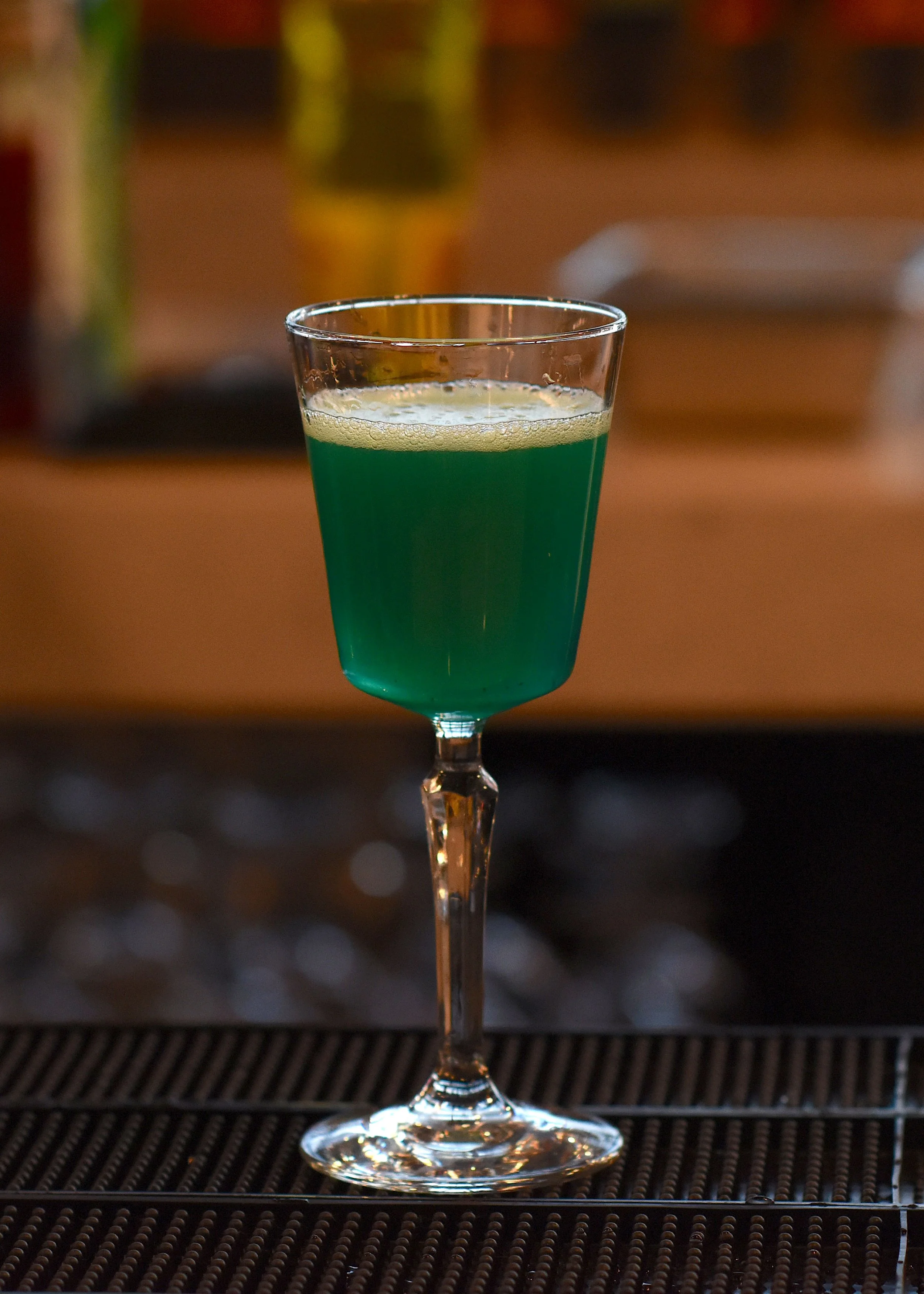 A glass with a long stem filled with a bright green cocktail, placed on a bar mat with a blurred background.