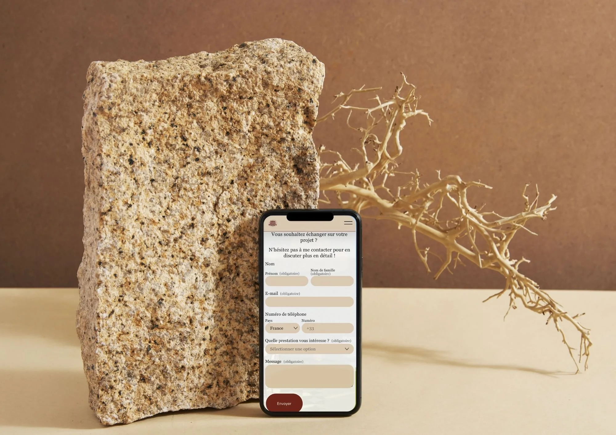 Téléphone affichant un formulaire de contact en français, posé devant une pierre et une plante sèche sur une table beige, avec un fond marron.