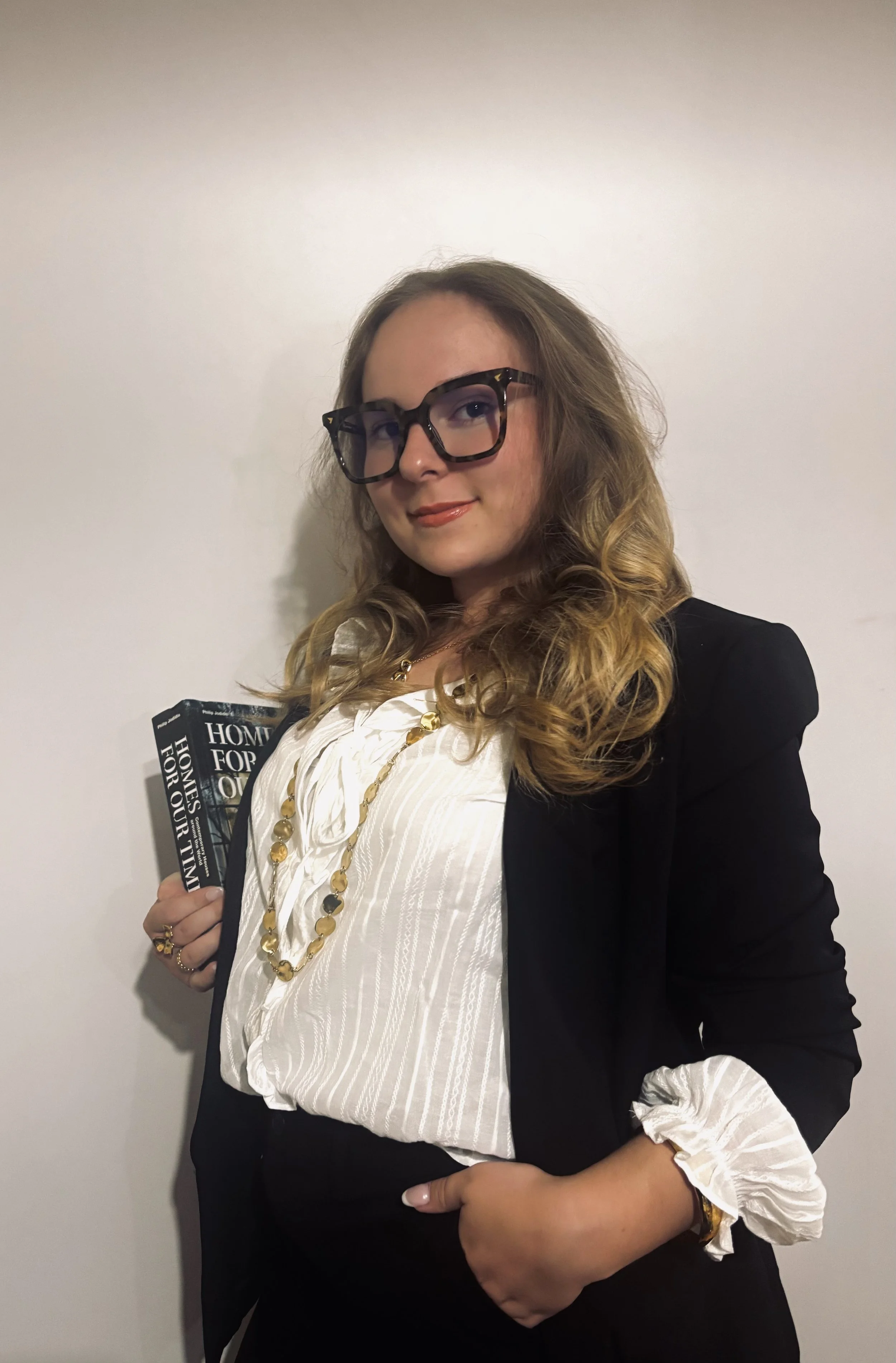 Une femme avec des lunettes épaisses, portant un blazer noir et une blouse blanche à manches froncées, tenant deux livres dans la main, devant un mur blanc.