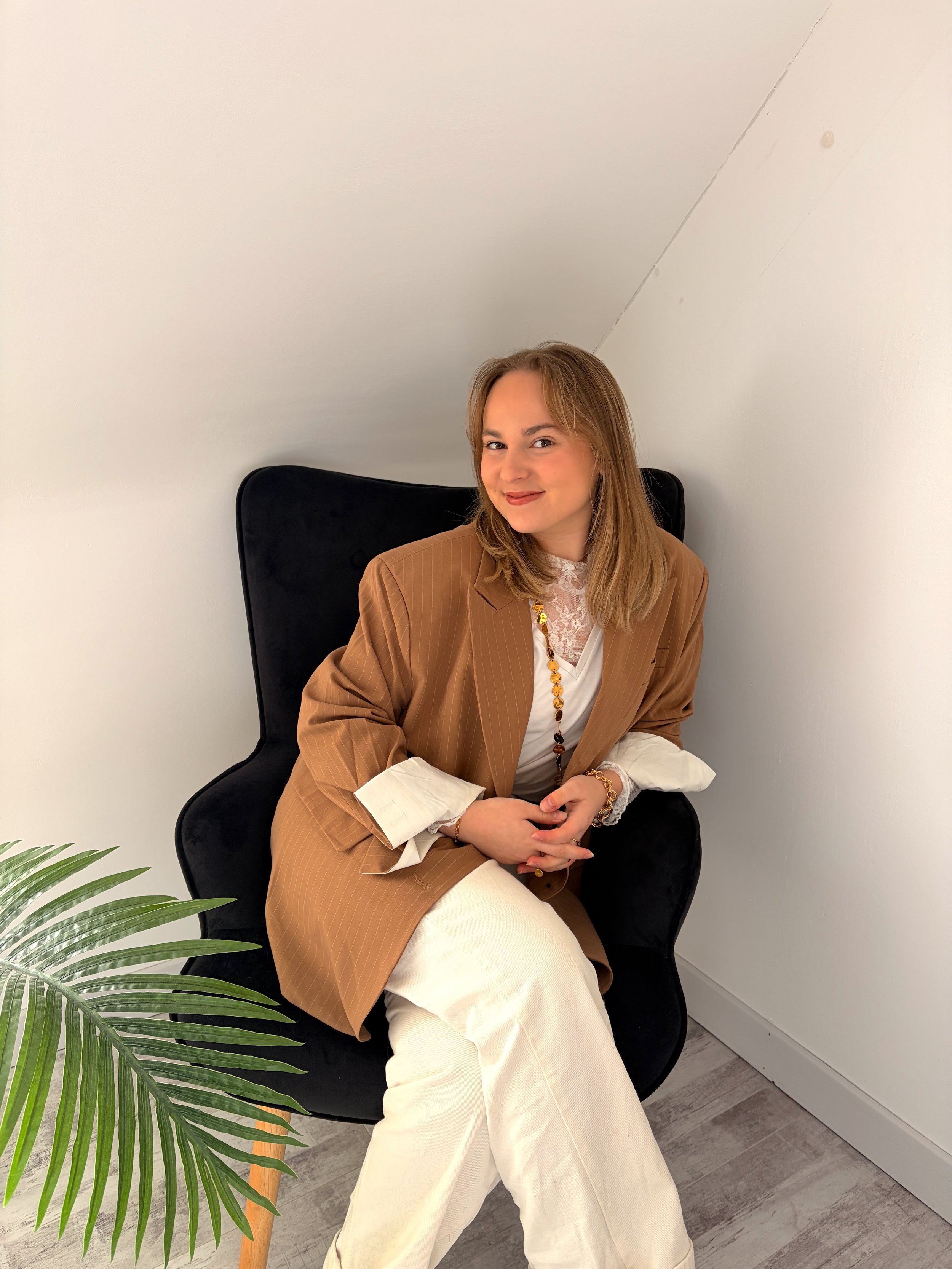 Femme assise sur une chaise noire, portant un blazer marron, une chemise blanche avec des détails en dentelle, un collier en perles, et des bracelets en or, avec un mur blanc en fond.