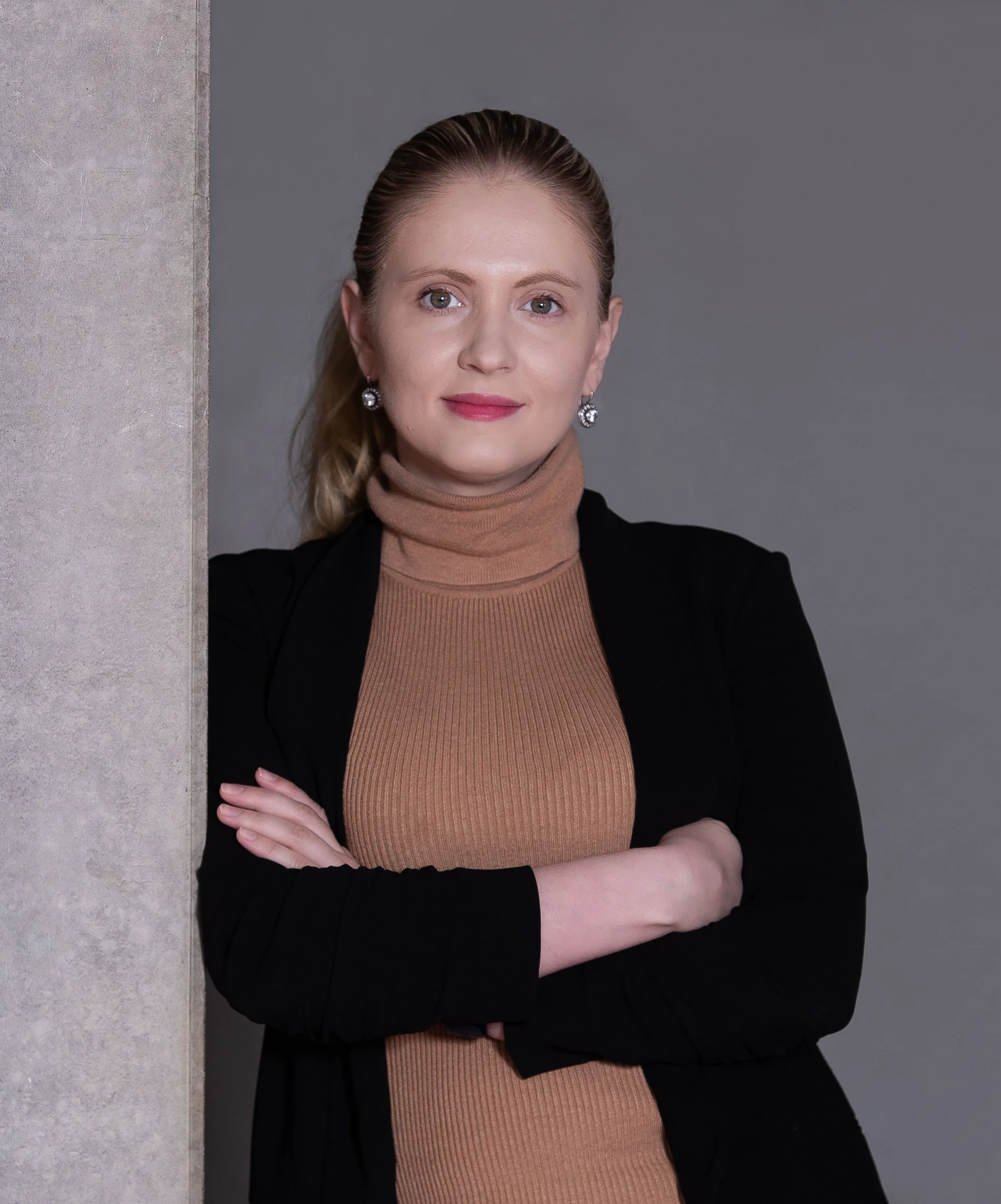 A woman with blonde hair, green eyes, wearing earrings, a tan turtleneck, and a black blazer, standing with arms crossed and leaning against a wall, looking at the camera.