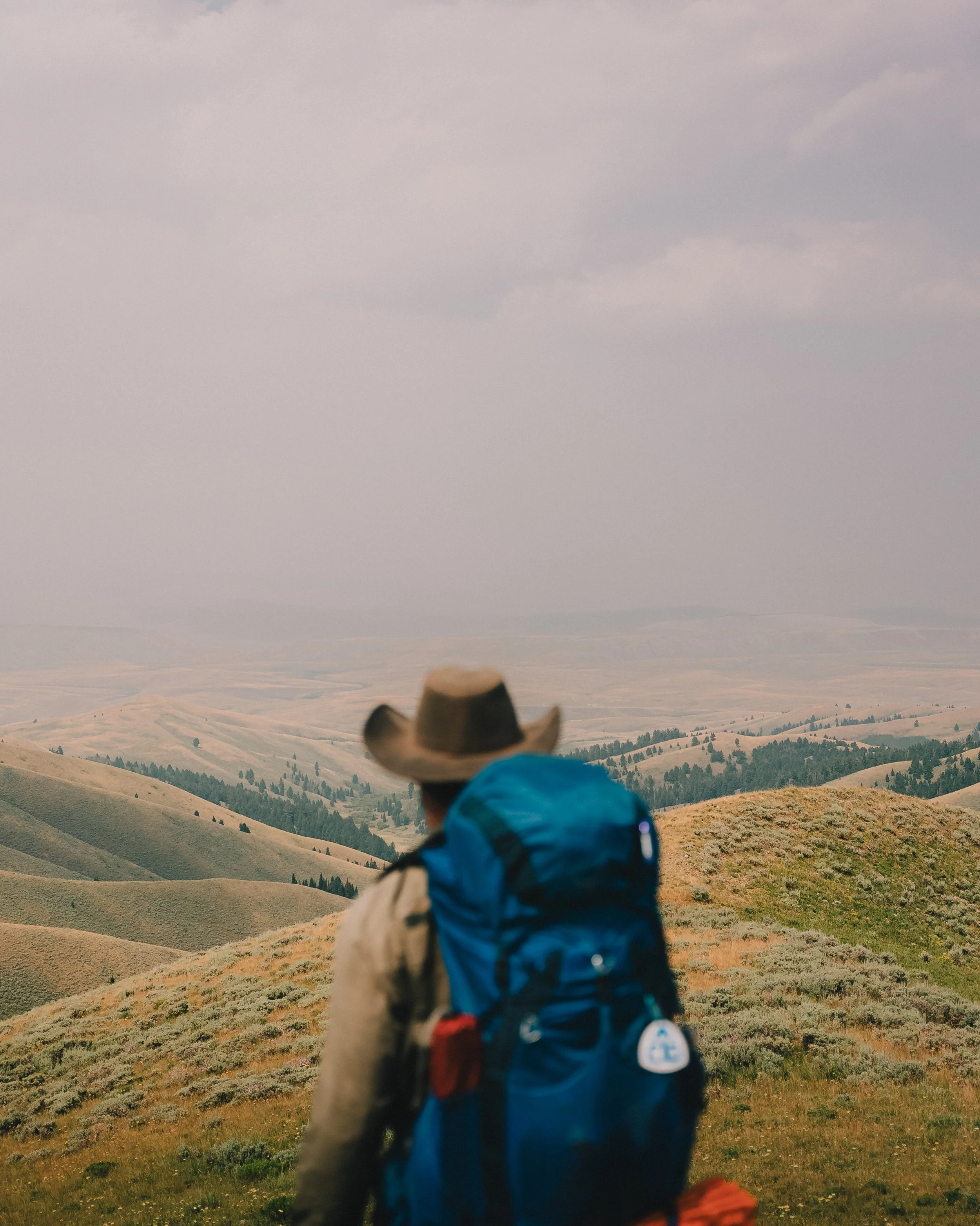 A person hiking in a hilly landscape wearing a cowboy hat and a blue backpack.