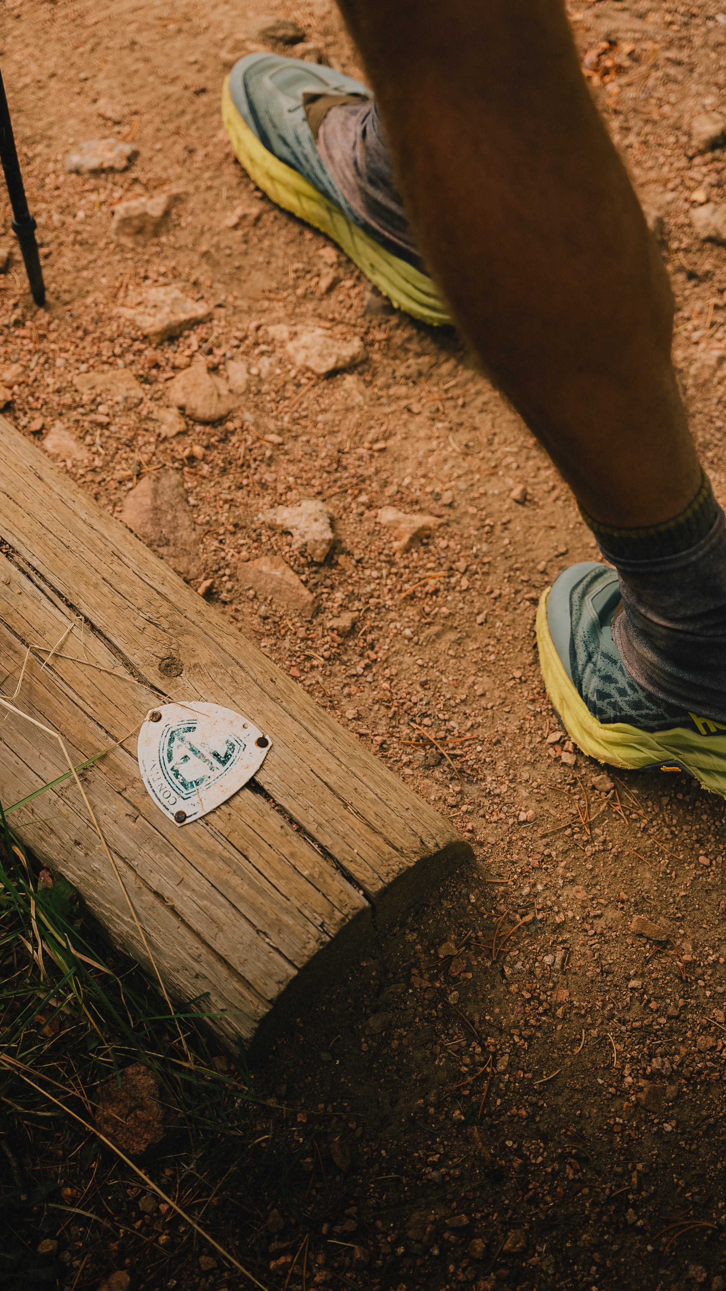 Close-up of a person's legs and feet wearing yellow running shoes and patterned socks on dirt trail with a wooden plank and a small trail marker.