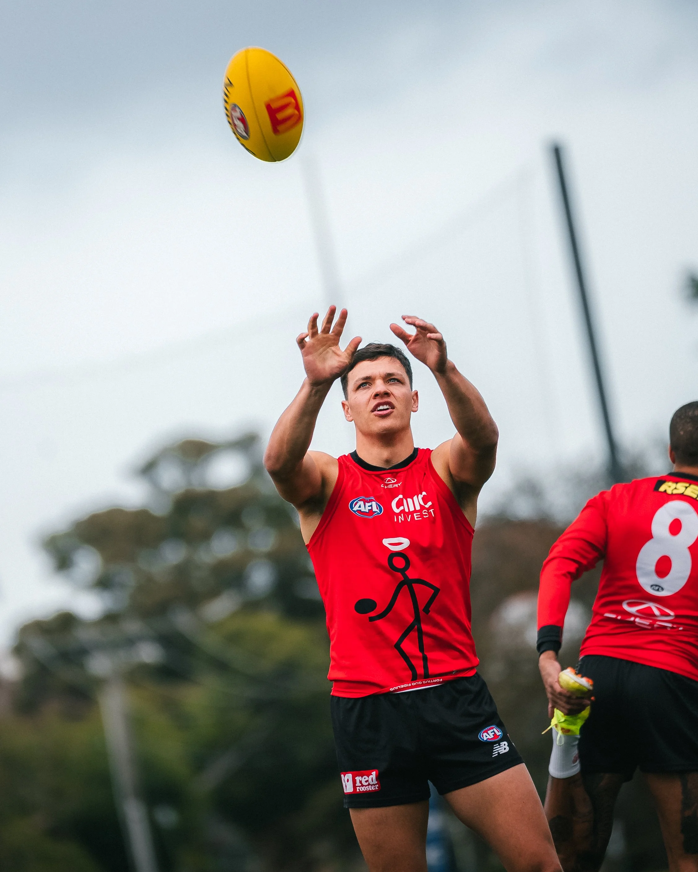Australian rules football player catching a ball during a game, with an overcast sky and trees in the background.